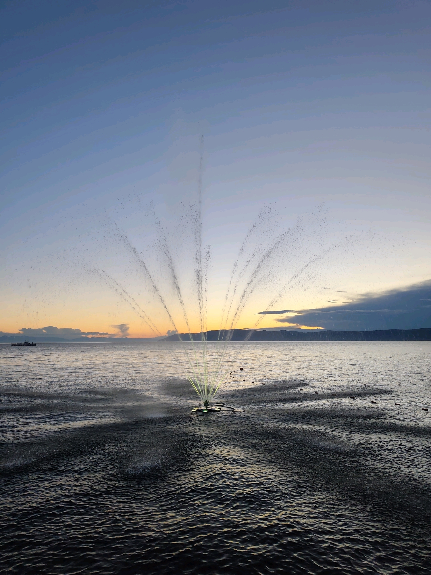At dusk, a fountain sprays water in a semi-circular pattern over the dark, rippling surface of a body of water, likely Lake Baikal, near Listvyanka, Russia. The sky transitions from a pale blue at the top to hues of orange and yellow near the horizon, where distant, hazy landmasses are visible. A few clouds drift above the horizon. In the distance, a small boat sails on the water. The water is disturbed by the fountain's spray, creating a visible pattern of ripples and splashes that extend outwards from the central device.