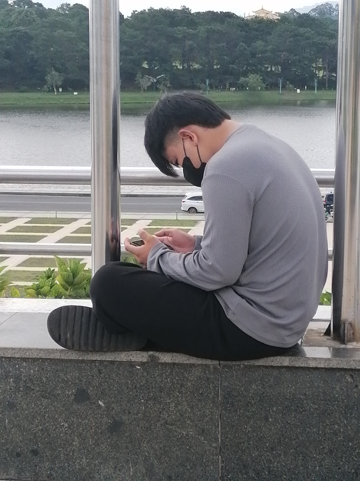 A young Vietnamese man wearing a grey long-sleeved shirt, black pants, and a black face mask is sitting on a grey stone ledge, looking down at his phone. He is in a relaxed, hunched position. Behind him, a body of water is visible, bordered by a line of trees and a building with a distinctive pagoda-style roof in the distance. A road with a white car and other vehicles is visible beyond the water, separated by a low railing. The man is positioned between two vertical metal poles that are part of a structure. The foreground consists of the grey stone ledge the man is sitting on. The scene appears to be in the Xuan Huong Lake area of Da Lat, Vietnam, known for its scenic views and recreational activities.