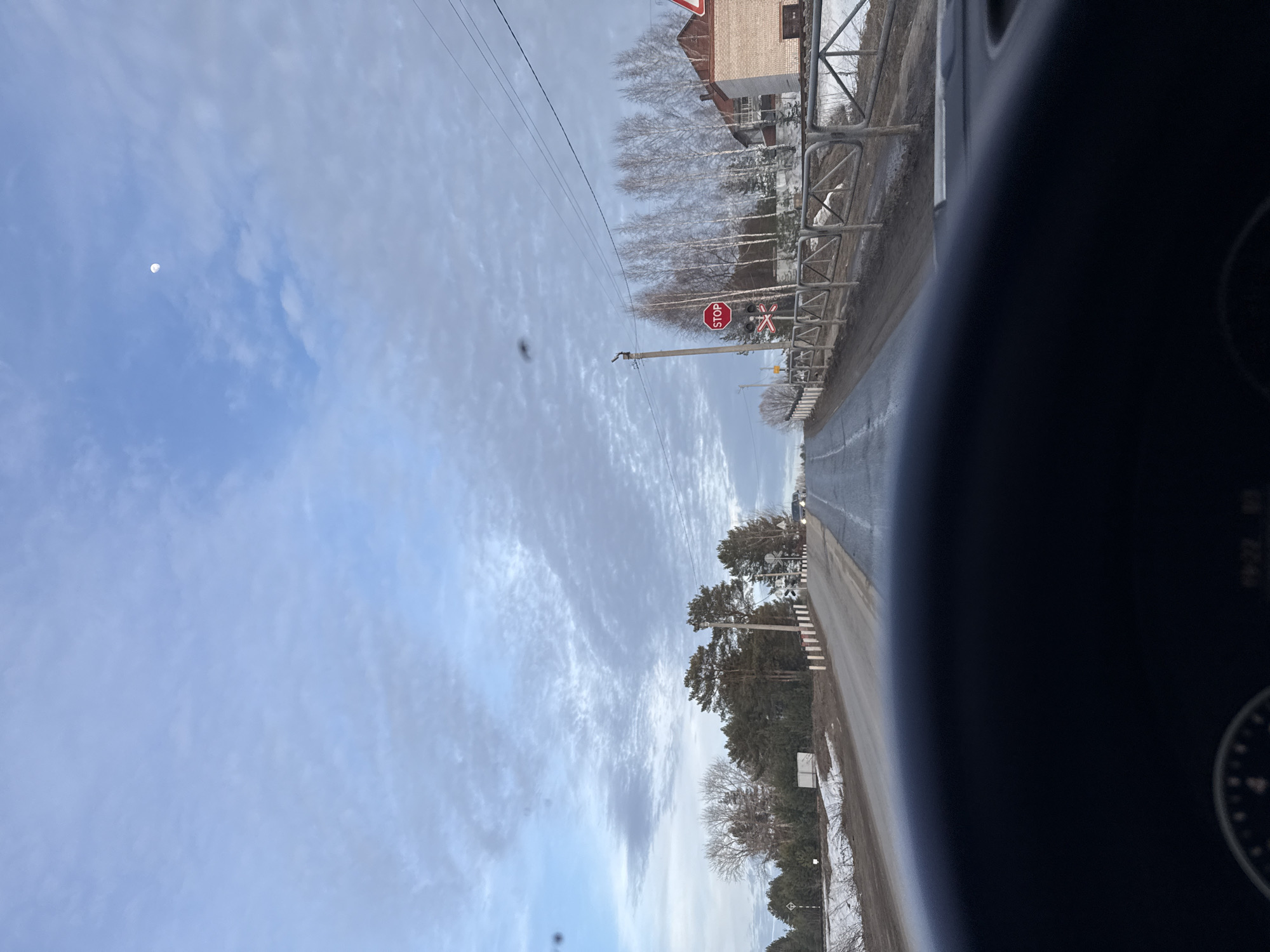 The image presents a forward-facing view from inside a vehicle, with the upper portion of a black steering wheel and a segment of the dashboard, displaying "19:22," visible in the lower foreground. The scene depicts a rural or semi-urban road, presumably in Chaikovsky, Russia, characterized by asphalt pavement with dirty shoulders and patches of snow.
The road ascends slightly and curves gently to the right in the mid-distance. On the left side, beyond the shoulder, there are bare deciduous trees and several evergreen pine trees, with a white diamond-shaped sign on a pole. A small, light-colored rectangular object, possibly a sign or barrier, is visible further back on the left shoulder.
On the right side of the road, a metal fence with an "X" pattern runs parallel to the asphalt. Behind this fence, several tall, slender, bare deciduous trees are visible. Further along this side, a two-story building constructed of light-colored brick with a reddish roof is partially visible. A red circular "STOP" sign is prominently displayed on a pole, accompanied by a white St. Andrew's Cross railway crossing sign directly below it. Another triangular warning sign with a red border is partially visible on the far right. Overhead power lines traverse the upper right quadrant of the image. Headlights from an oncoming vehicle are discernible in the distance on the right side of the road.
The sky is largely covered by stratocumulus clouds, with areas of lighter blue visible. A crescent moon is discernible in the upper right portion of the sky. A small, dark speck, likely dust or a smudge on the windshield, is present in the upper central area of the frame. No individuals are clearly identifiable.