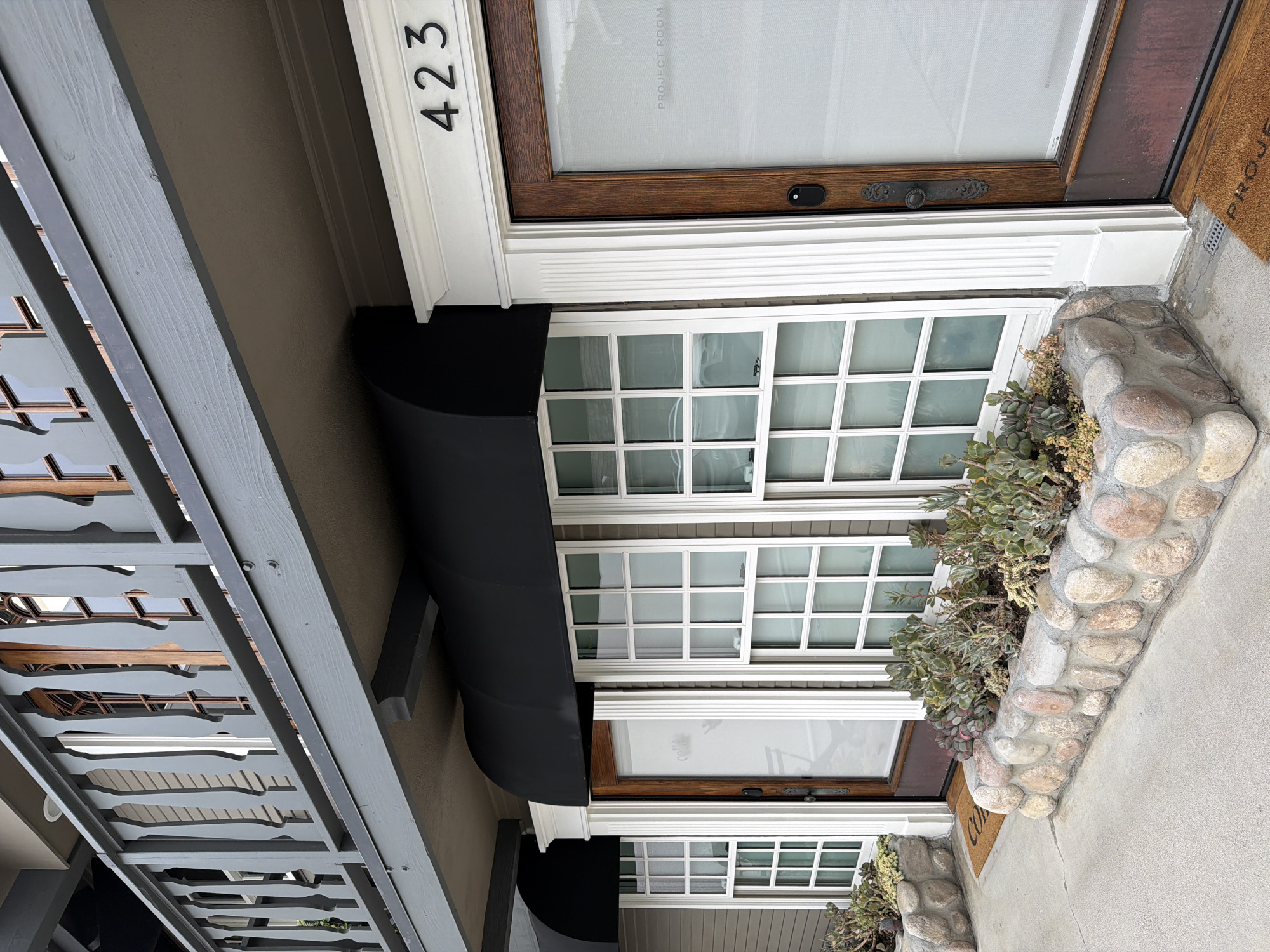 The image captures the exterior of a building located at 423 in Newport Beach, California. The architecture features a front door and adjacent windows with white grilles. A black awning extends over the windows. Stone landscaping is present near the building's entrance. A grey, decorative porch roof or balcony overhangs the entrance area. The photograph appears to be taken during daylight hours.