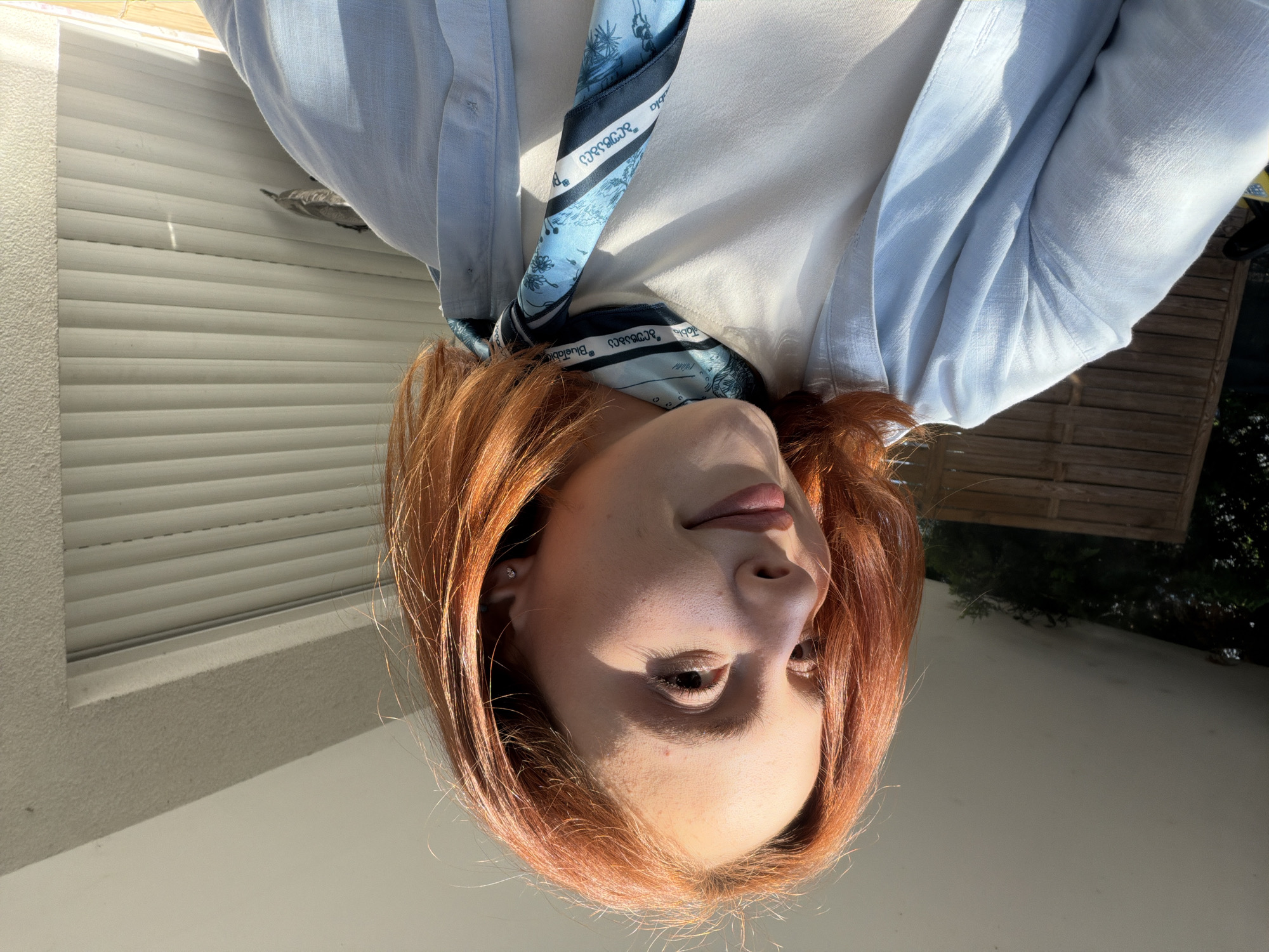 The image features a young adult female with medium-length, reddish-orange hair, smiling gently and looking towards her right (viewer's left). Strong natural sunlight illuminates her face and hair, creating distinct highlights and shadows. She is wearing a white crew-neck t-shirt layered under an open, light blue, textured long-sleeved shirt. Around her neck is a light and dark blue patterned scarf, adorned with flora designs and text including "BlueTable," "בלוּטֵבּל" (Hebrew for Blue Table), and "ცისფერი მაგიდა" (Georgian for Blue Table).
The environment is an outdoor residential setting, consistent with a balcony or patio in Strasbourg, France. To the left of the frame, a smooth, light gray ceiling or overhang is visible above a wooden slatted fence, behind which green foliage and trees can be seen. To the right, a textured light gray wall segment rises above a closed, horizontally slatted roller shutter, a common architectural feature in the Alsace region. The strong, low-angle sunlight suggests either morning or late afternoon.