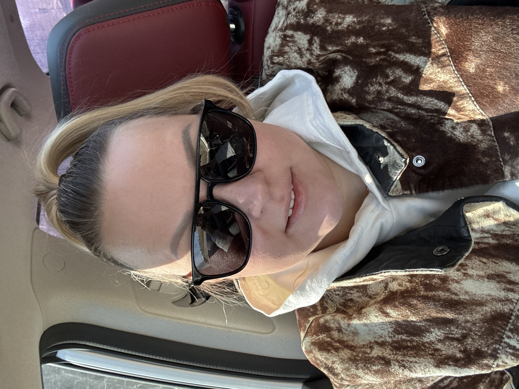 A single adult female is depicted in a chest-level, head-on photograph taken inside a vehicle, located in the city of Хохряки, Russia. The subject has blonde hair, with darker roots, pulled into a high ponytail. She is wearing dark, rectangular sunglasses, obscuring her eyes, and a subtle, closed-lip smile is visible. Her attire includes a light-colored hooded sweatshirt beneath an outer jacket. The jacket exhibits a distinct mottled pattern of dark brown and light cream/white, resembling cowhide or a similar fur texture, with black leather accents and silver snap buttons visible on the collar area. She is seated, likely in a front passenger position, with a red leather car seat featuring black stitching visible behind her head. The vehicle's interior ceiling and left pillar are light-toned. Strong, natural light illuminates the subject from the left, creating defined shadows.