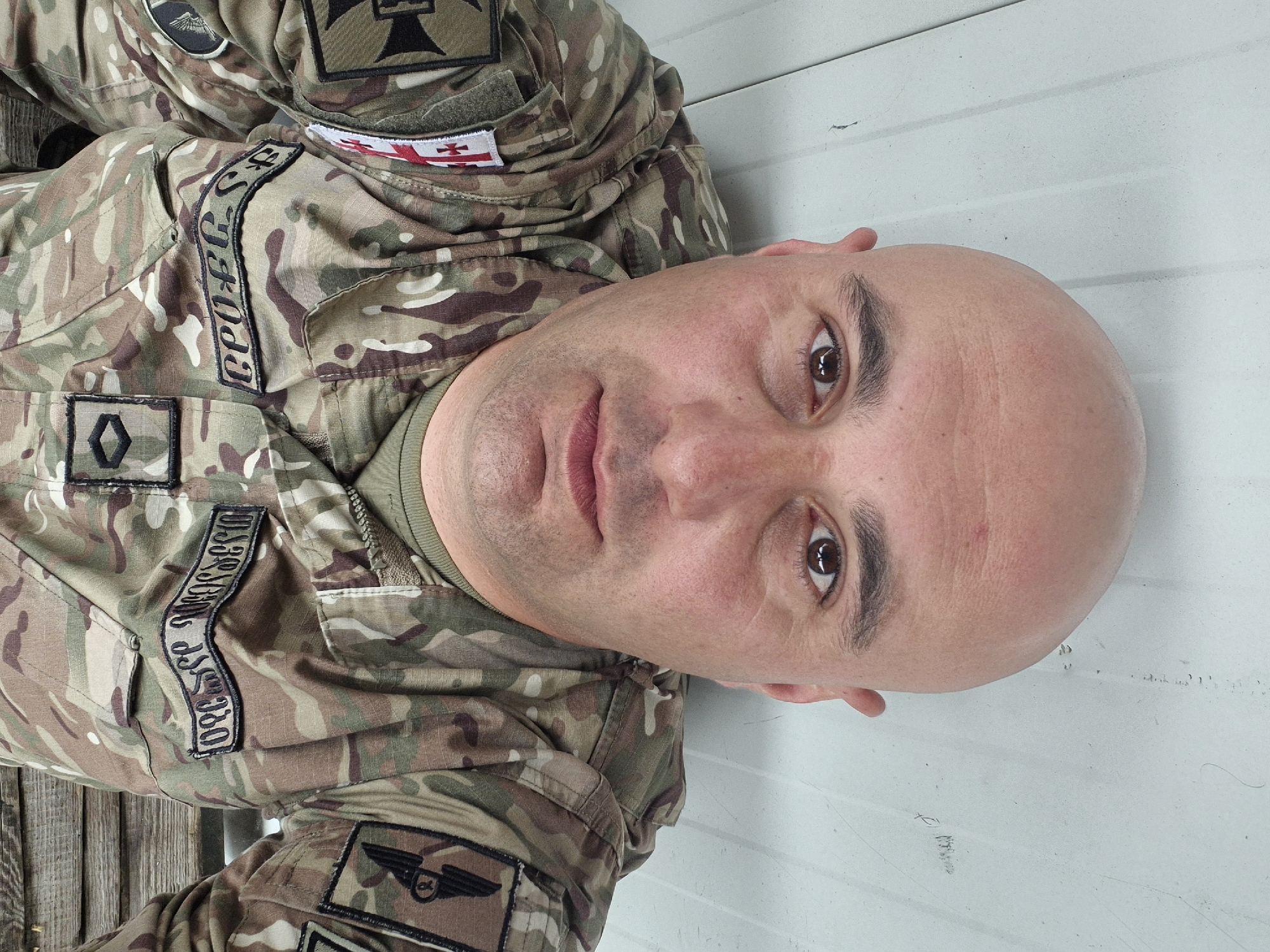 The image is a close-up, eye-level shot of a bald, light-skinned man wearing a camouflage military uniform. The uniform has patches indicating Georgian affiliation, including the Georgian flag and name tag in Georgian script. He appears to be indoors, with a white wall visible in the background. The location in Tbilisi, Georgia, is not directly discernible from the image.