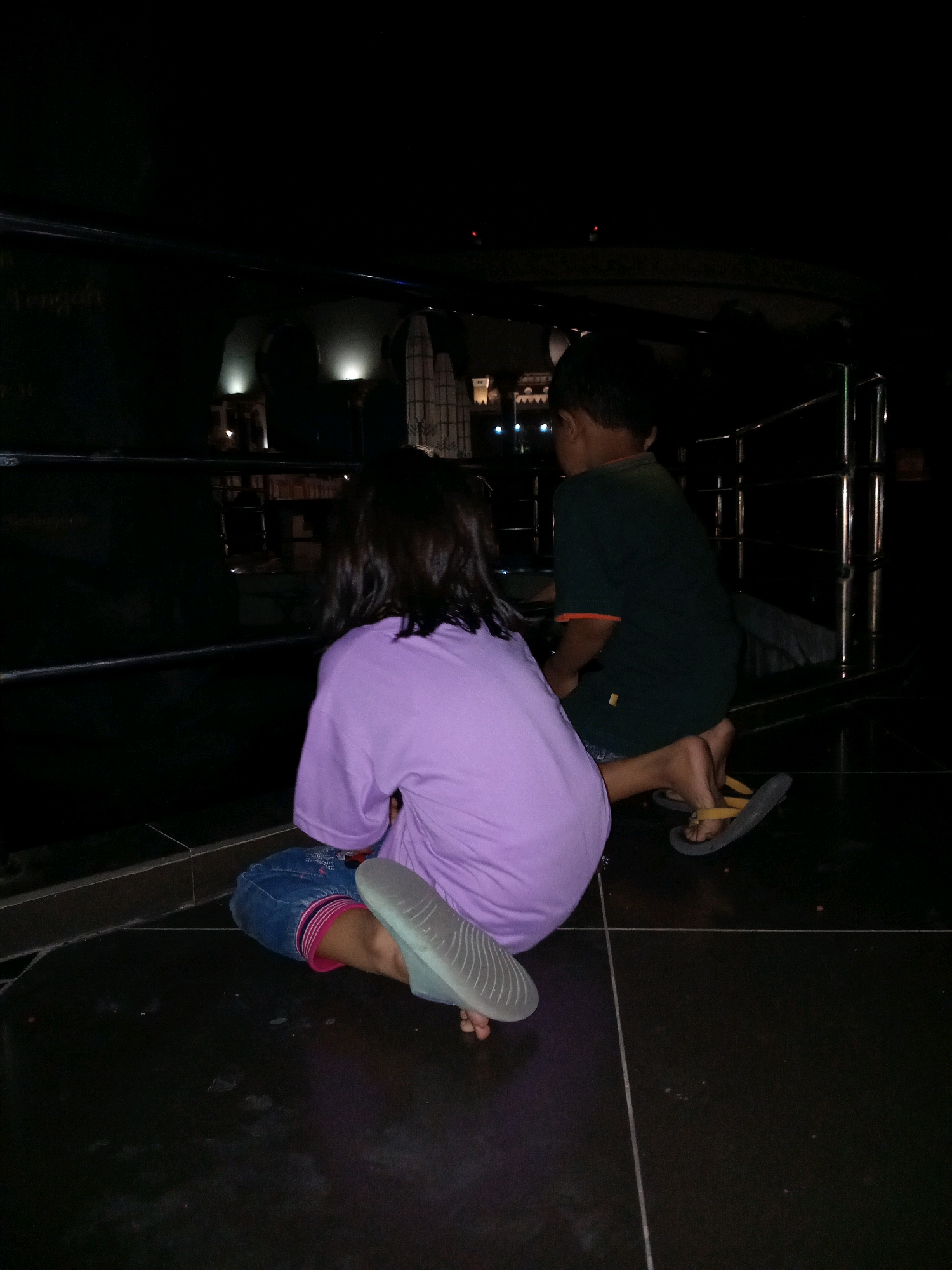 In Semarang, Indonesia, two children are seen from behind in a dark environment, possibly an indoor public space with dimly lit architectural elements in the background. The child on the left is wearing a purple t-shirt and denim shorts with pink trim, and appears to be wearing light blue sandals. The child on the right is wearing a dark green t-shirt with an orange trim and appears to be wearing dark flip-flops. Both children are kneeling on a dark tiled floor. A metal railing is visible in the background, suggesting a walkway or elevated area. The overall lighting is dim, with artificial lights illuminating some distant structures.