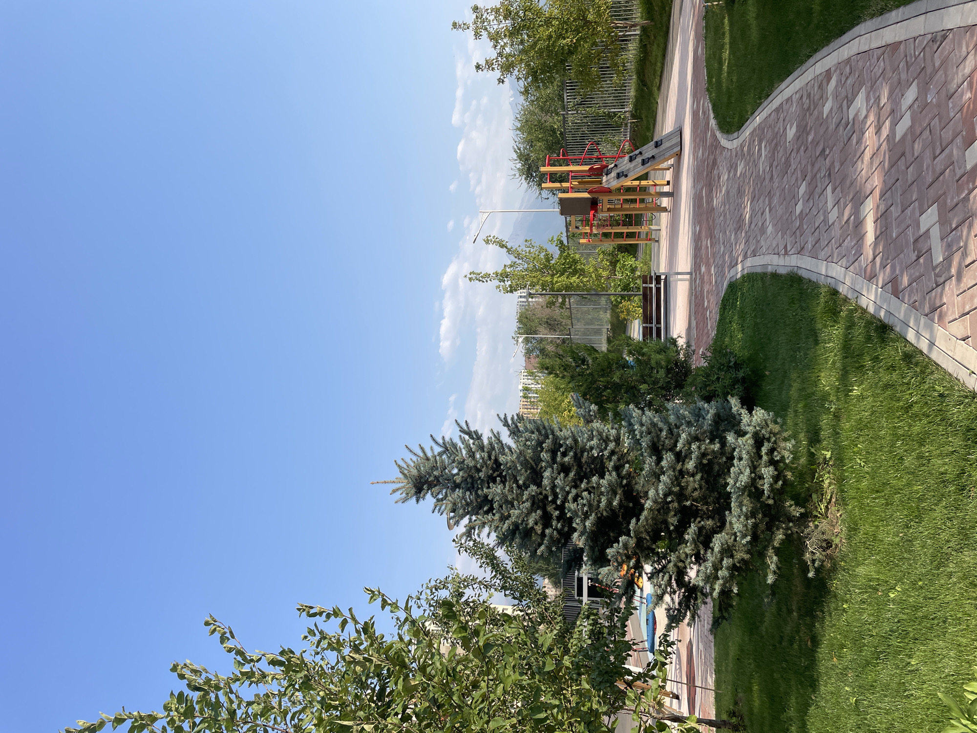 An outdoor scene features a playground and a pathway in the city of Almaty, Kazakhstan. The pathway, made of red and beige interlocking bricks, curves through a grassy area. To the left, a large tree with green leaves stands beside a blue-colored splash pad or shallow pool. In the background, a playground structure with red and yellow elements is visible. A metal fence encloses the playground and separates it from trees and distant buildings. Mountains and a bright blue sky with scattered white clouds form the backdrop. The scene appears to be a public park or residential courtyard on a sunny day.