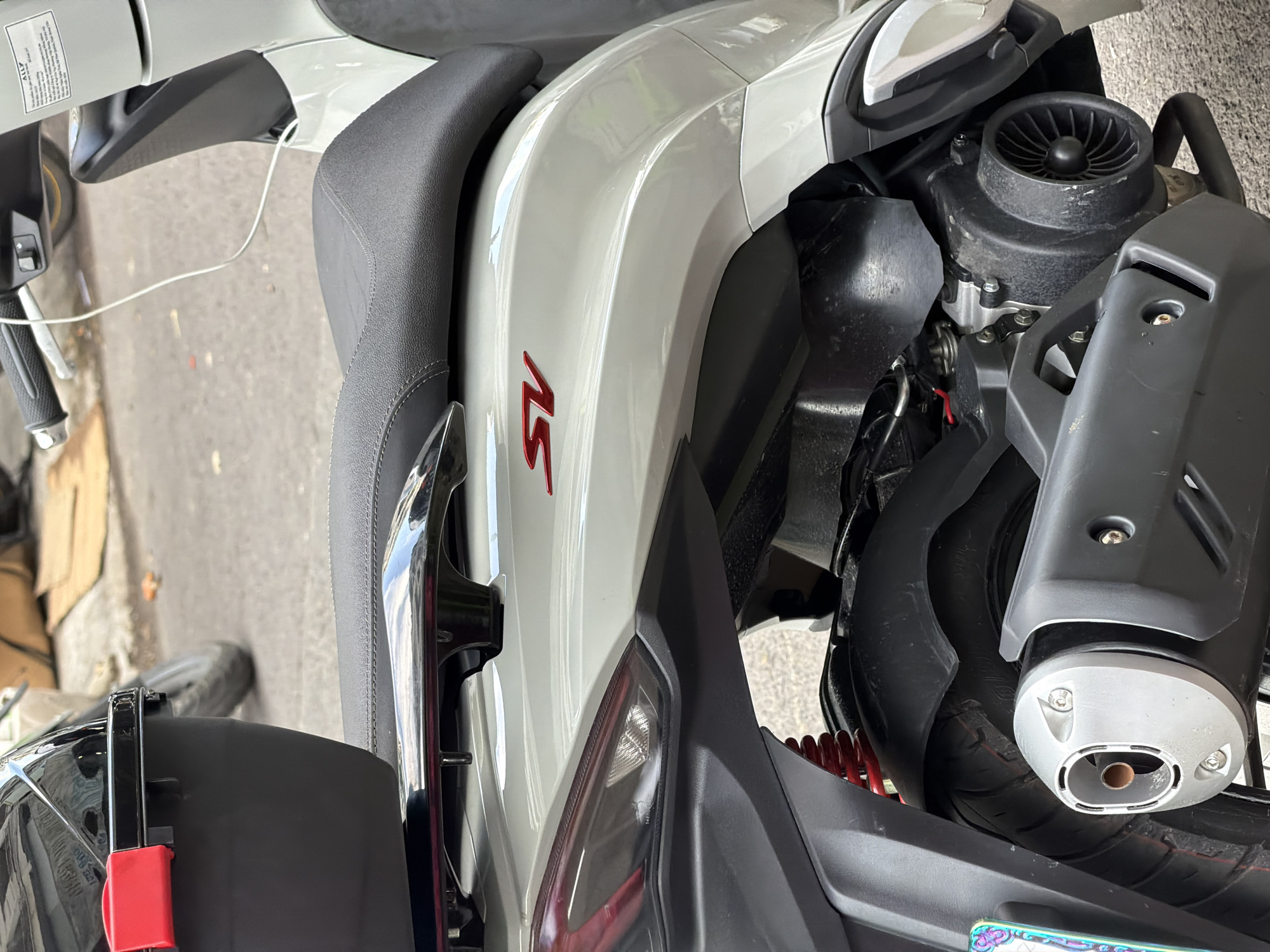 The image presents a close-up, high-angle view of a white scooter, parked outdoors on a paved surface, likely a street or sidewalk, in Nha Trang Ward, Vietnam. The scooter's right side is prominently featured, illuminated by bright daylight, suggesting a clear and possibly sunny day.
The primary subject is the scooter itself, displaying a glossy white body contrasted with a black textured seat and various dark gray and black plastic components. A notable detail is the red "SV" emblem or text affixed to the scooter's white side panel, just below the seat.
Towards the rear of the scooter, mechanical elements are visible, including a black engine cover with a fan-like structure, a red coiled spring indicating a rear shock absorber, and a silver-capped exhaust pipe leading from the rear wheel assembly. A sleek, chrome-like grab handle extends from the back of the seat.
On the far left, a portion of the handlebar with a black grip and a brake lever is visible, along with a thin white cable descending towards the ground. A white sticker near the handlebars bears the text "ALLY" above some unreadable smaller text. At the bottom left, a red strap or clip, possibly part of a helmet restraint, is attached to a black fixture. A small, colorful segment of a license plate or sticker is also visible at the very bottom right of the frame. No people or specific activities are depicted; the scooter appears to be stationary.