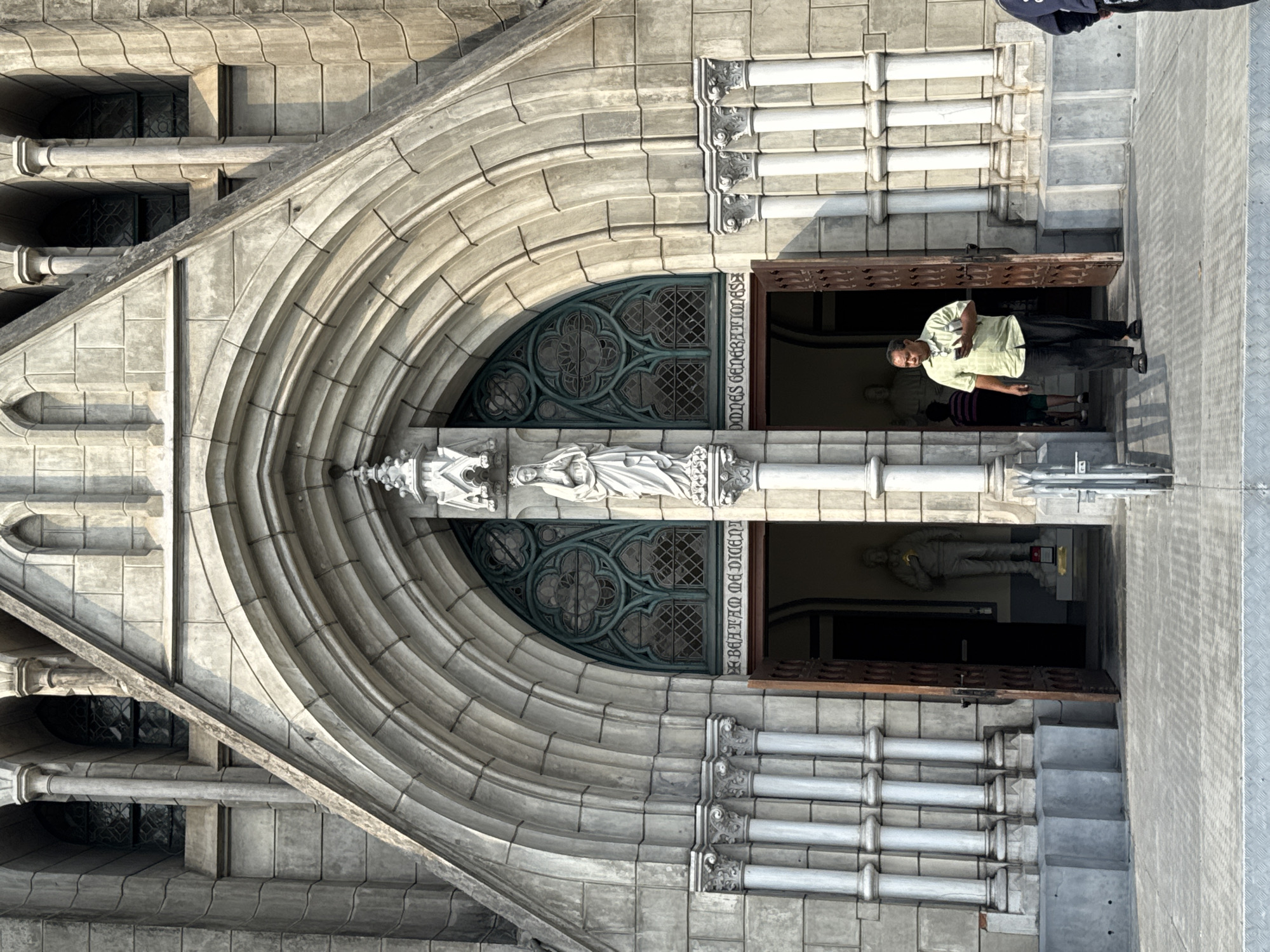The image shows the entrance to the Jakarta Cathedral, a Roman Catholic cathedral in Central Jakarta, Indonesia. The architecture is Gothic Revival style, featuring a large arched doorway with intricate stonework and a statue of the Virgin Mary above the entrance. A man stands in the doorway, appearing to be either entering or exiting the cathedral. The environment suggests a bright, sunny day. The photograph is taken from a slightly low angle, emphasizing the height and grandeur of the building. Text is visible on the side of the door frame.