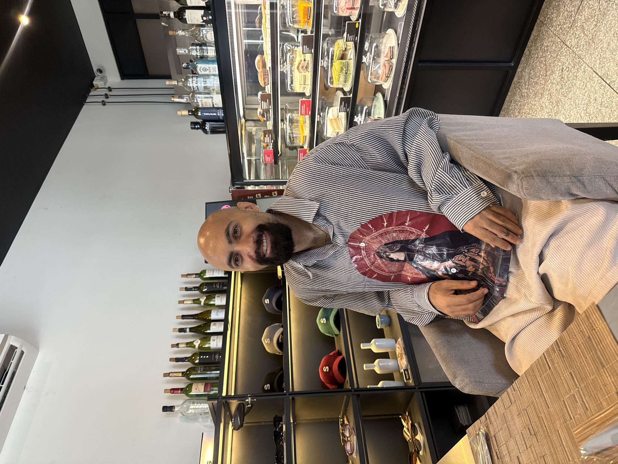 A bald man with a beard sits in a gray chair in what appears to be a cafe or shop. He is wearing a striped shirt with a graphic print on the front and light-colored pants. The man is smiling and looking at the camera. Behind him, shelves are stocked with bottles of alcohol and hats. A display case filled with cakes and pastries is visible to the right. The man's hands are resting on his lap, and a small tattoo is visible on his left hand. The overall environment suggests a retail establishment with a seating area.