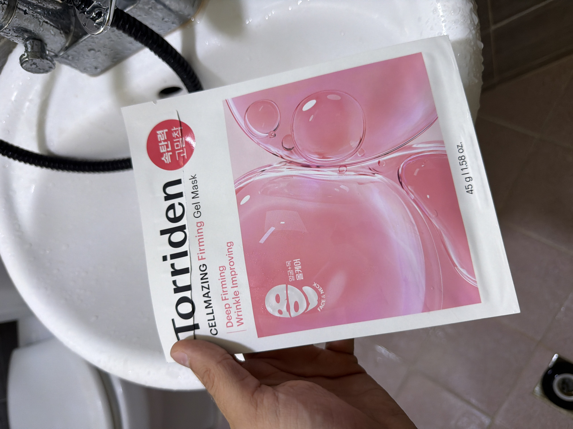 An image shows a person's hand holding a Torriden CELLMAZING Firming Gel Mask in a bathroom. The mask packaging is pink and white with text indicating it is for "Deep Firming Wrinkle Improving" and "Face & Neck" care. A red circular emblem on the packaging contains Korean text. The background is a white sink with water droplets on its surface and a chrome faucet. The floor tiles are brown. There is no indication of the location being in Seoul, South Korea.