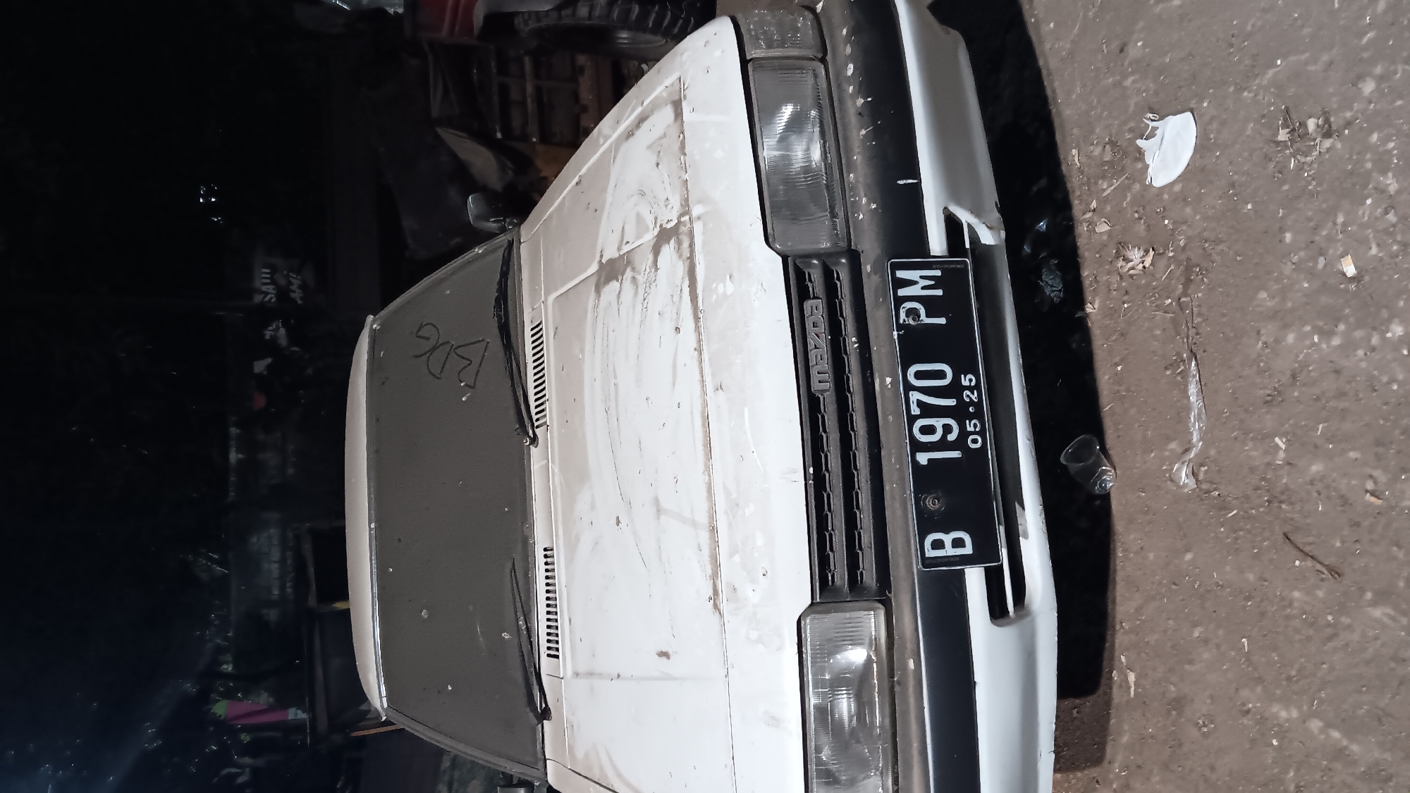 The image shows the front of a white, dusty Mazda sedan parked outdoors at night. The car has a license plate that reads "B 1970 PM 05-25". The grille of the car has the "Mazda" logo. The windshield has writing on it that appears to be "BDG". There is debris on the ground in front of the car, including a small glass and some plastic wrappers. The background is dark and shows trees and structures, suggesting an urban or semi-urban environment. The scene does not explicitly indicate a location in South Jakarta, Indonesia, but the license plate format "B" for Jakarta plates and the general appearance are consistent with an Indonesian setting.