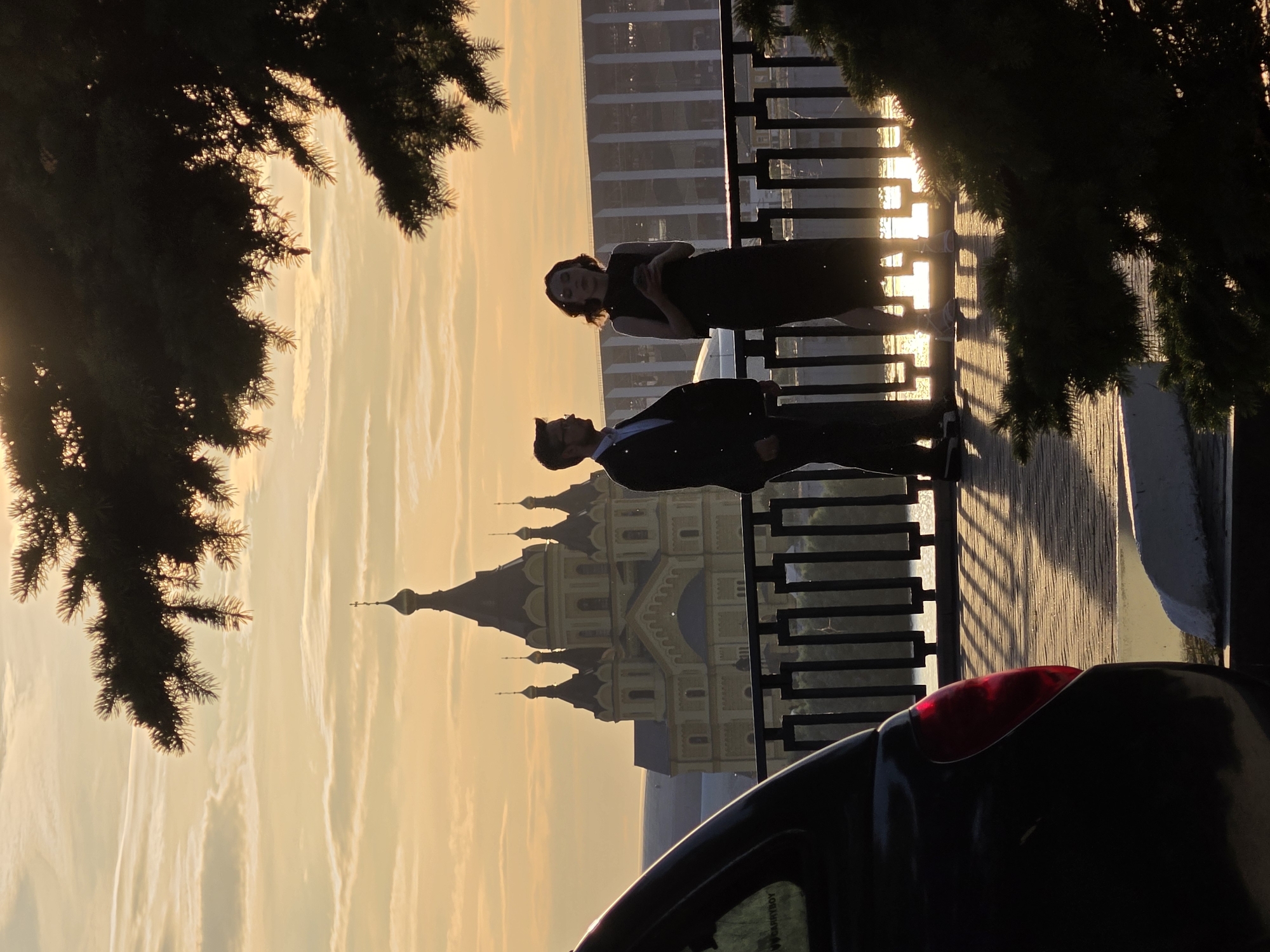 The photograph captures a sunset scene in Nizhny Novgorod, Russia, featuring the Nizhny Novgorod Kremlin in the background. In the foreground, a black car is partially visible on the left. Two individuals, a man in a suit and a woman in a dark dress, stand near a railing, silhouetted against the golden sky. The man is looking to his left, while the woman is looking down at her phone. Behind them, the architectural grandeur of the Kremlin, with its distinctive domes and spires, is illuminated by the setting sun. The environment appears to be an elevated viewpoint, possibly a park or promenade, with a body of water visible below the railing. The scene is framed by the dark branches of a coniferous tree in the upper right corner. The overall atmosphere is one of serene beauty, enhanced by the warm hues of the sunset.