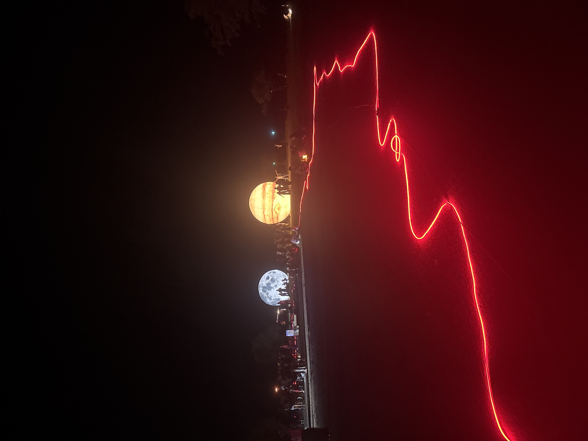 The image depicts an outdoor nighttime scene in Baan Pang Ka, Thailand, featuring two large, illuminated inflatable spheres resembling planets or moons. The sphere on the left is textured to look like the moon, while the one on the right is yellow with stripes, possibly representing Jupiter. Numerous people are gathered around these illuminated objects, some standing and others sitting. In the foreground, a bright red neon light rope snakes across the ground, creating a winding path. The ground appears to be a dark, somewhat sandy or gravelly surface. The background is dark, indicating nighttime, with some trees visible on the right side. The overall atmosphere suggests an art installation or event taking place outdoors.