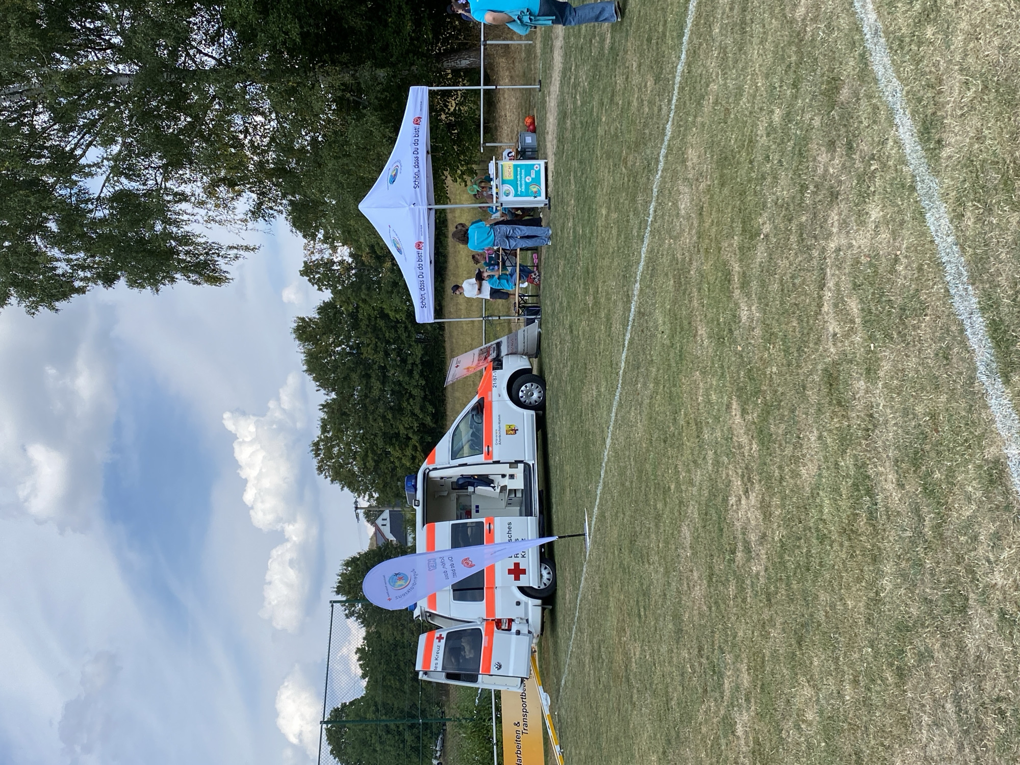An outdoor scene on a dry grass field, possibly a sports field marked with white lines, under a partly cloudy sky. A white ambulance, identifiable by its red cross emblems and emergency lights, is parked to the left. The ambulance doors are open, revealing its interior. A white event tent with a banner that reads "Schön, dass Du da bist!" (Beautiful that you are here!) is set up to the right. Underneath the tent, several people, including children and adults, are gathered around tables. A white flag with a teardrop shape stands in front of the ambulance. Trees line the background, and a chain-link fence is visible further back. The overall atmosphere suggests a community event or gathering in Ingelbach, Germany.