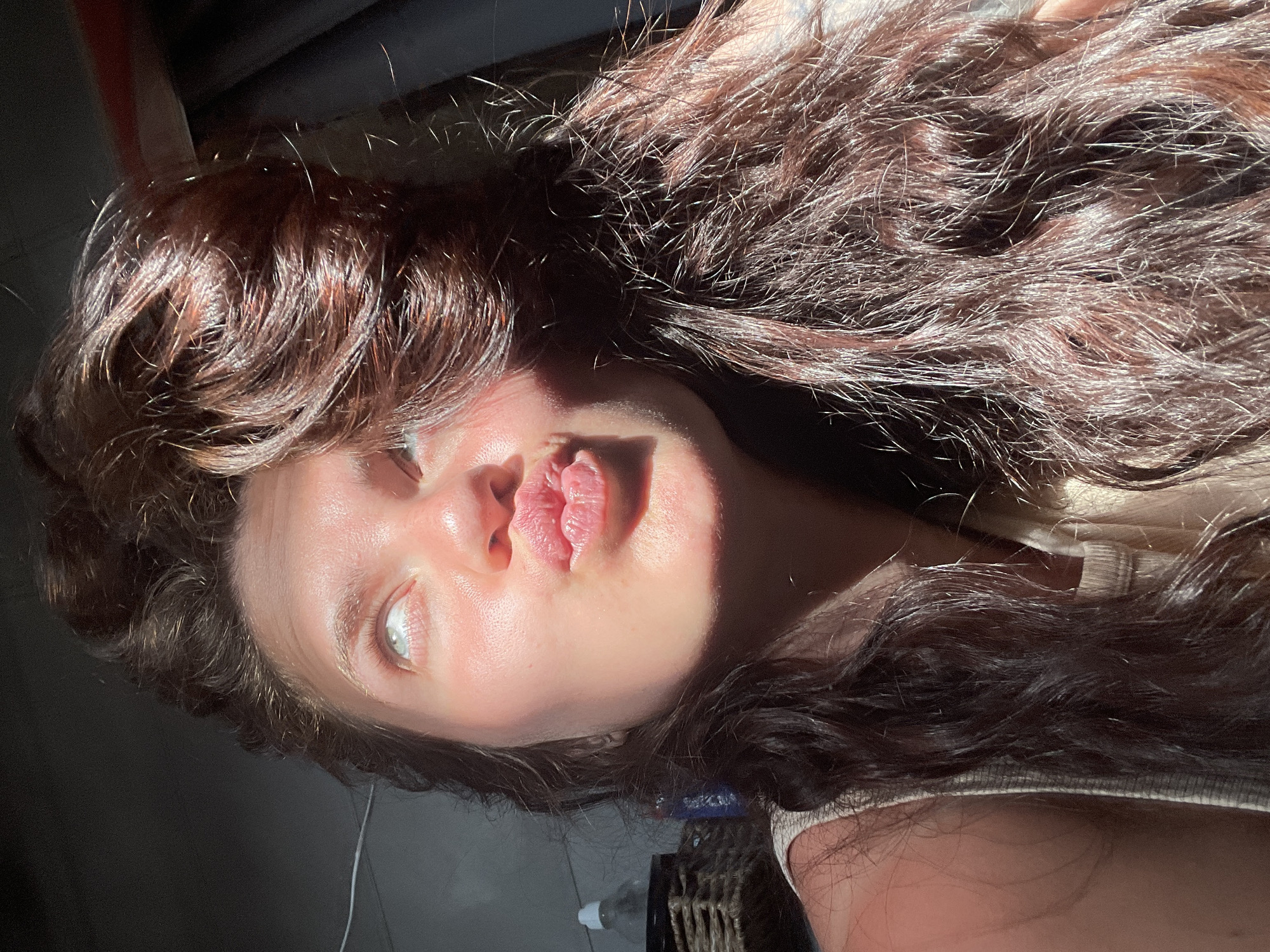A close-up shot captures a young woman with long, dark, wavy hair making a kissing face towards the camera. Sunlight dramatically illuminates the left side of her face, casting shadows and highlighting the texture of her skin and hair. Her eyes are looking upwards and slightly to the left, with her lips pursed. She is wearing a light-colored sleeveless top. In the background, a blurred basket and a bottle are visible on the left, and a dark curtain or wall on the right. The scene appears to be indoors, possibly in a home or studio, with natural light streaming in. The location of Мосметаллоконструкция Онт, Russia is not determinable from the image.
