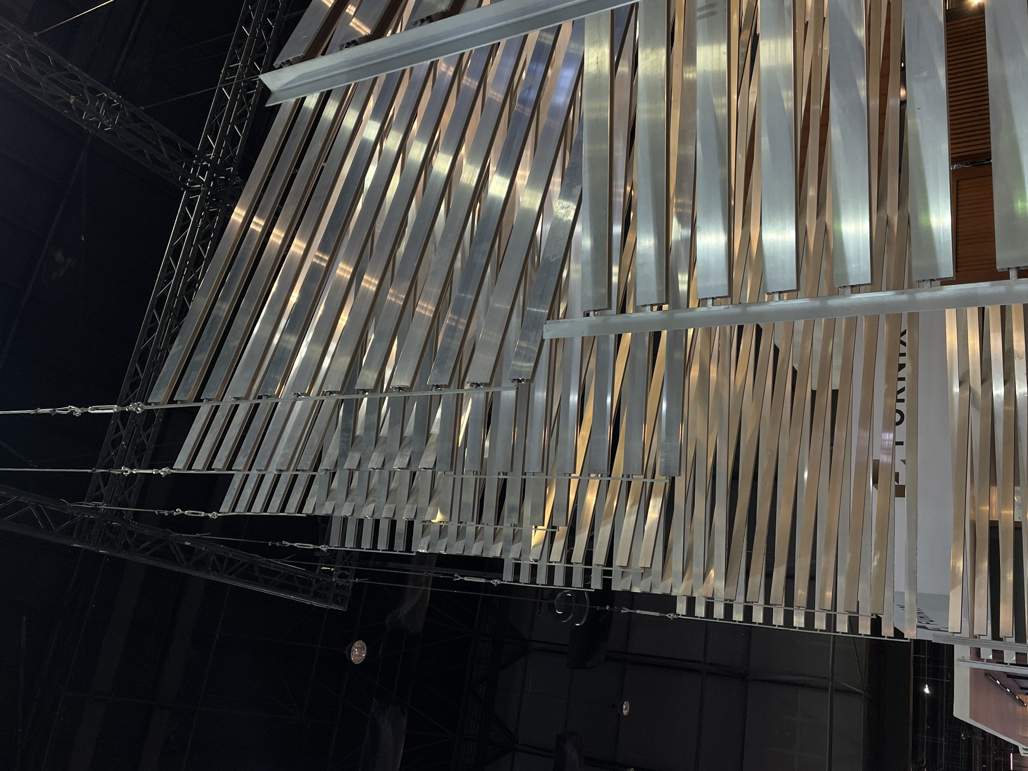 The image shows an abstract art installation inside an event space in Pak Kret City Municipality, Thailand. Multiple reflective metal strips hang vertically from overhead supports, creating a dynamic visual effect with light and shadow. The strips are of varying lengths. Visible above is a metal framework, and below is a dark background with lighting fixtures. A wall is also visible in the bottom right-hand corner of the image.