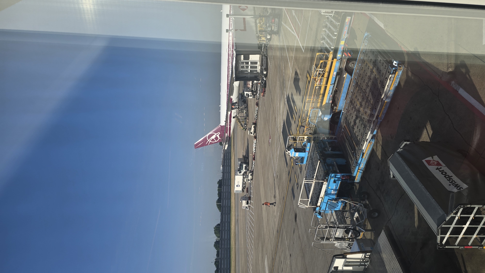 The image depicts an airport scene, likely taken from inside a building through a glass window. An airplane with a maroon tail fin is visible on the tarmac, connected to a jet bridge. Ground support equipment, including luggage carts and vehicles, is positioned around the plane. One person wearing an orange safety vest is visible on the tarmac. The equipment has the brand "Swissport" on the side. Reflections on the window obscure the scene and the sky. The geographical location within the city of London, United Kingdom, is not conclusively determined from the visual information provided.