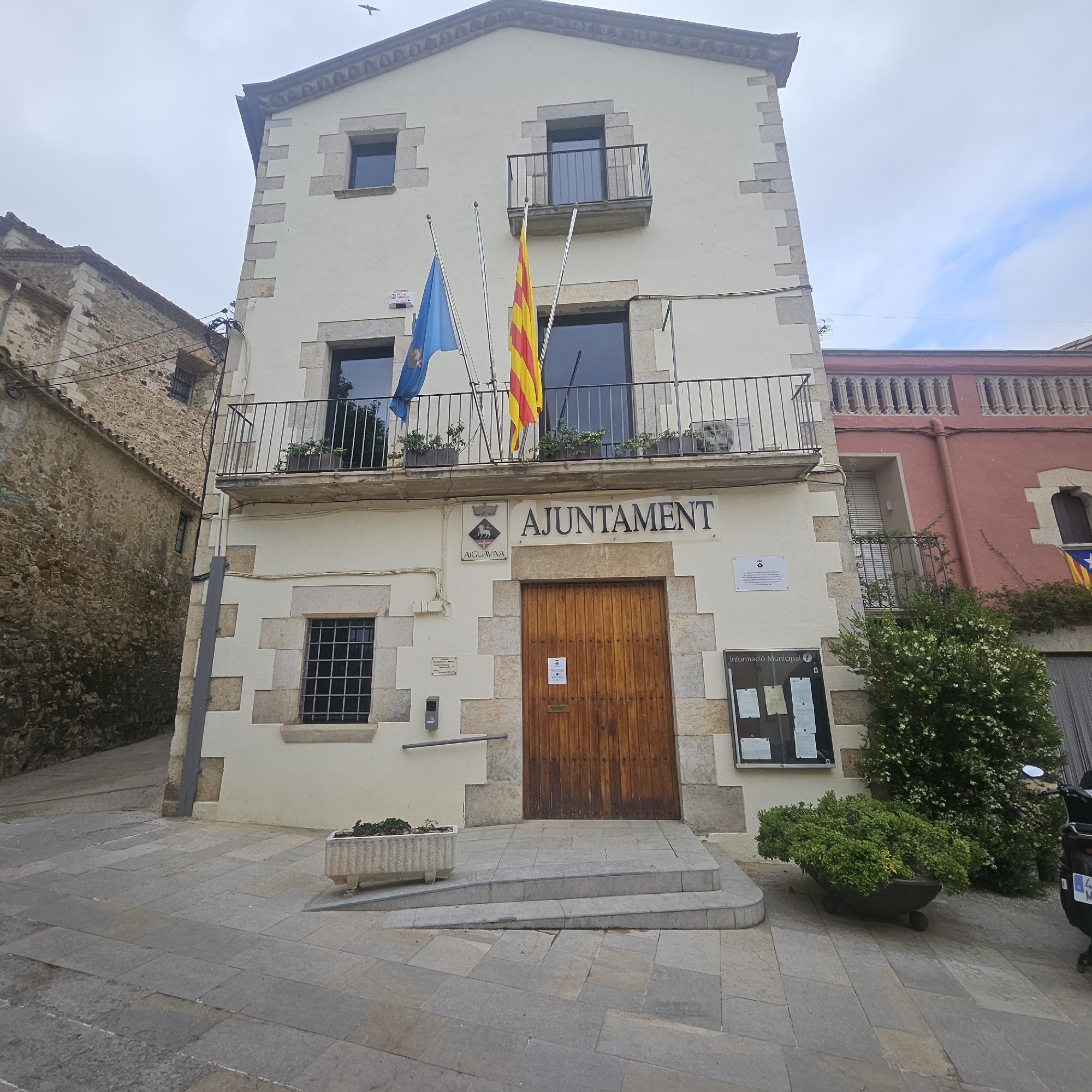 The image shows the Ajuntament (town hall) building in Aiguaviva, Spain. The building is a light-colored, multi-story structure with stone accents. Two flags, one blue with an emblem and another yellow and red striped, fly from poles on the second story balcony. Above the entrance doors, the word "AJUNTAMENT" is prominently displayed. The entrance is a large wooden door, and a ramp with steps leads up to it. A planter with green foliage is located to the left of the entrance. Adjacent to the Ajuntament, a stone building with a dark tiled roof is visible. To the right, a pink building with decorative window arches and another balcony with foliage is present. The ground is paved with large, flat stones. A motorcycle is parked on the right side of the frame. The sky is overcast with some blue patches and clouds, and a bird is visible flying in the upper portion of the image.