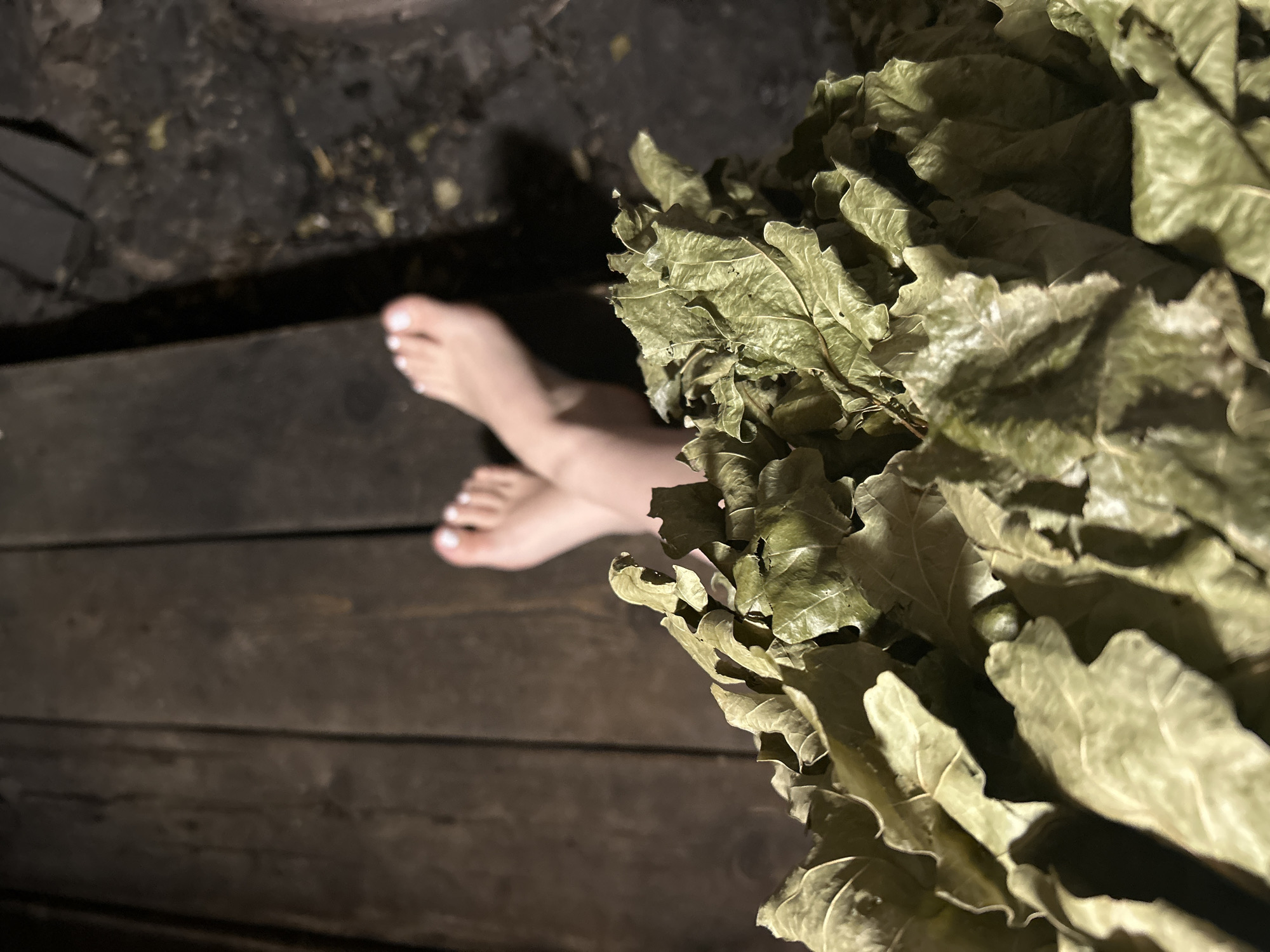 A dimly lit scene in what appears to be a sauna or bathhouse setting. A wooden floor stretches horizontally, and a woman's feet, painted with white nail polish, are visible in the middle-left. To the right, a bundle of dried birch leaves is prominently displayed. The exact location is unspecified, but based on the prompt, it's likely situated in the city of Chaikovsky, Russia. No other people are visible, and the interaction is static, with the feet resting beside the birch leaves. The environment is dark and rustic, indicative of a traditional bathing space.