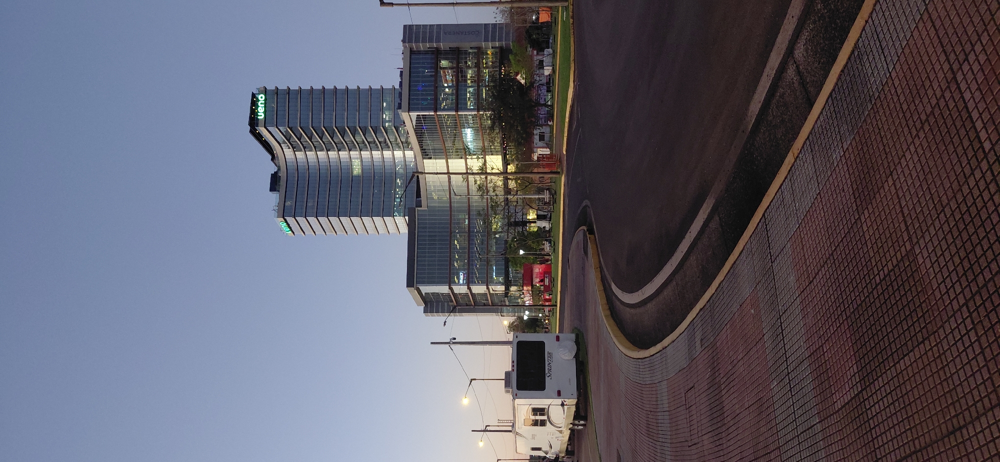 The image captures a view of a modern building with the "ueno" logo illuminated in green at its apex, situated in Encarnacion, Paraguay. The building, which appears to be a corporate office or a mixed-use complex, features a curved, multi-story glass facade reflecting the clear sky and surrounding lights. In the foreground, a paved sidewalk with a red and gray patterned brick surface lines a curved asphalt road. A white recreational vehicle, identified as a "Sprinter," is parked on the sidewalk to the left of the frame. Streetlights are visible, casting a soft glow on the scene, and trees line the area behind the sidewalk. The overall ambiance suggests an urban setting during twilight or early evening.