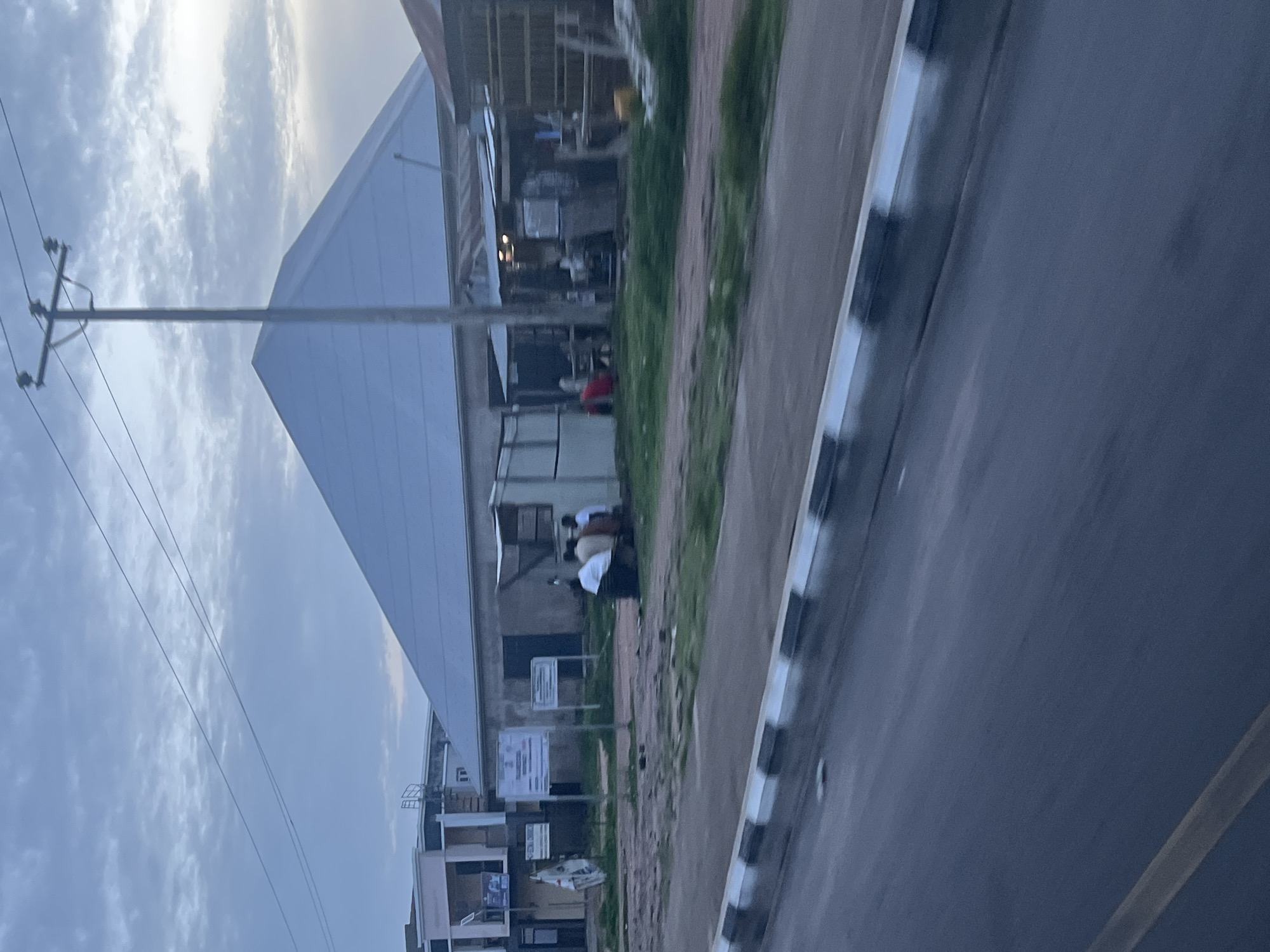 The image captures a street scene in Jalingo, Nigeria, likely during the late afternoon or early evening. The foreground features a paved road with a yellow line marking, and a distinct curb with black and white stripes. Beyond the road, a grassy area with scattered dirt patches separates the road from a collection of structures.
Dominating the midground is a large, pyramid-shaped building with a light-colored, possibly white or silver, roof. The sides of this structure appear to be made of concrete or a similar material, with some visible scaffolding or supports. To the left of this building, a two-story structure with a light-colored facade and windows is visible. Several signs are posted in front of this building, one appearing to be a noticeboard with text.
To the right of the pyramid-shaped building, there are several smaller, open-air structures with corrugated metal roofs. These appear to be shops or stalls, with some people visible inside and in front of them. One of these stalls has a light-colored wall and a dark opening. Further to the right, another structure with a corrugated metal roof and wooden supports is visible.
In front of the pyramid-shaped building and between it and the stalls, a group of people are gathered. They are seated or standing, and some are wearing white garments. The overall impression is a bustling, yet somewhat unrefined, urban environment with a mix of formal and informal architecture. A utility pole with wires stretches across the upper part of the image, passing in front of the pyramid-shaped roof. The sky above is overcast with clouds.