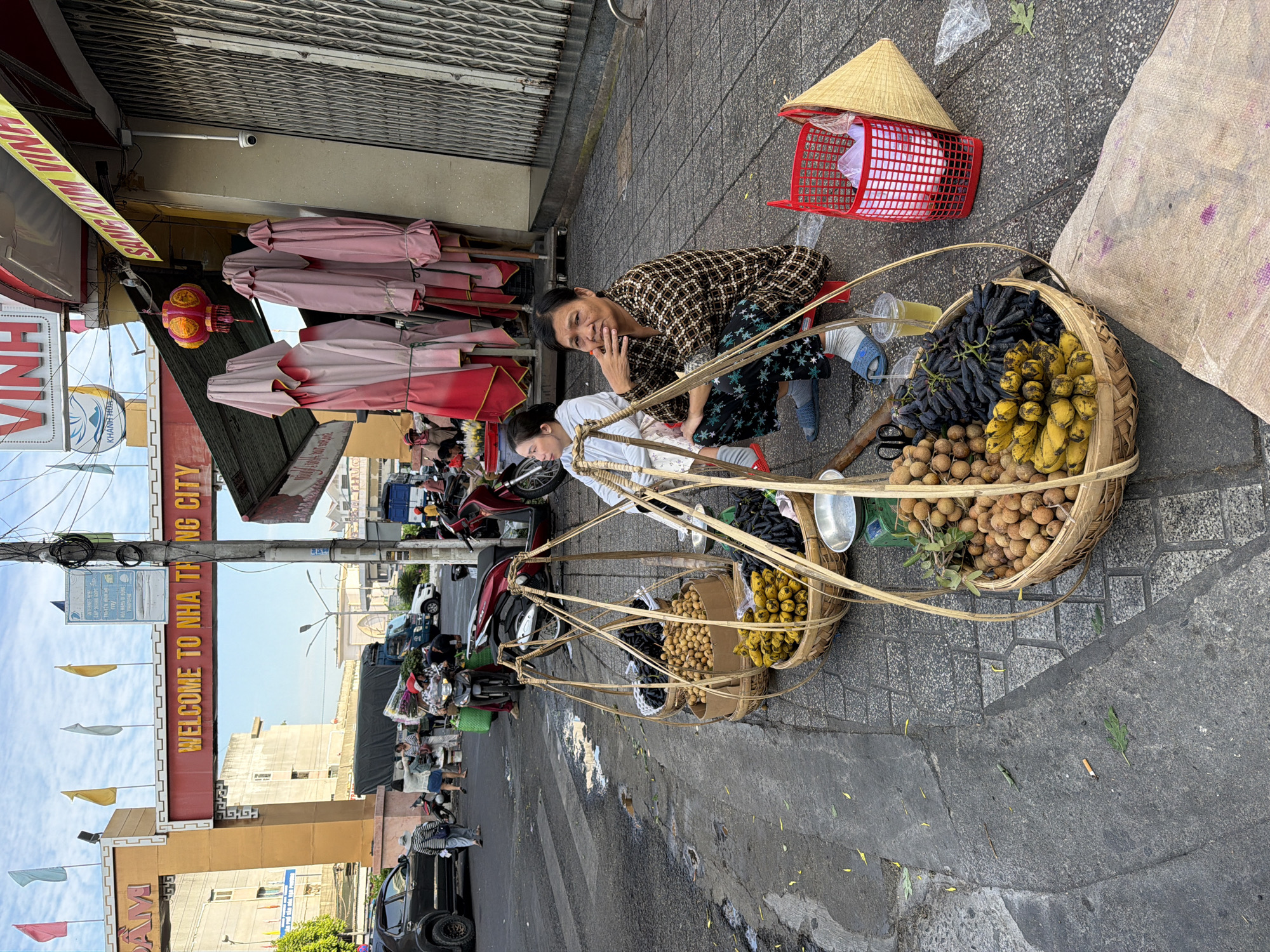 The image shows a street scene in Nha Trang, Vietnam. Two women are selling fruit from baskets on the sidewalk. The baskets are made of bamboo and contain bananas, longan, and dark-colored grapes. One woman is squatting next to the baskets, and the other is sitting behind them. In the background, there is an archway with the words "WELCOME TO NHA TRANG CITY" written on it. There are also shops and buildings, along with motorcycles and cars on the street. The sky is visible above, with some clouds. The overall impression is that of a busy street market.