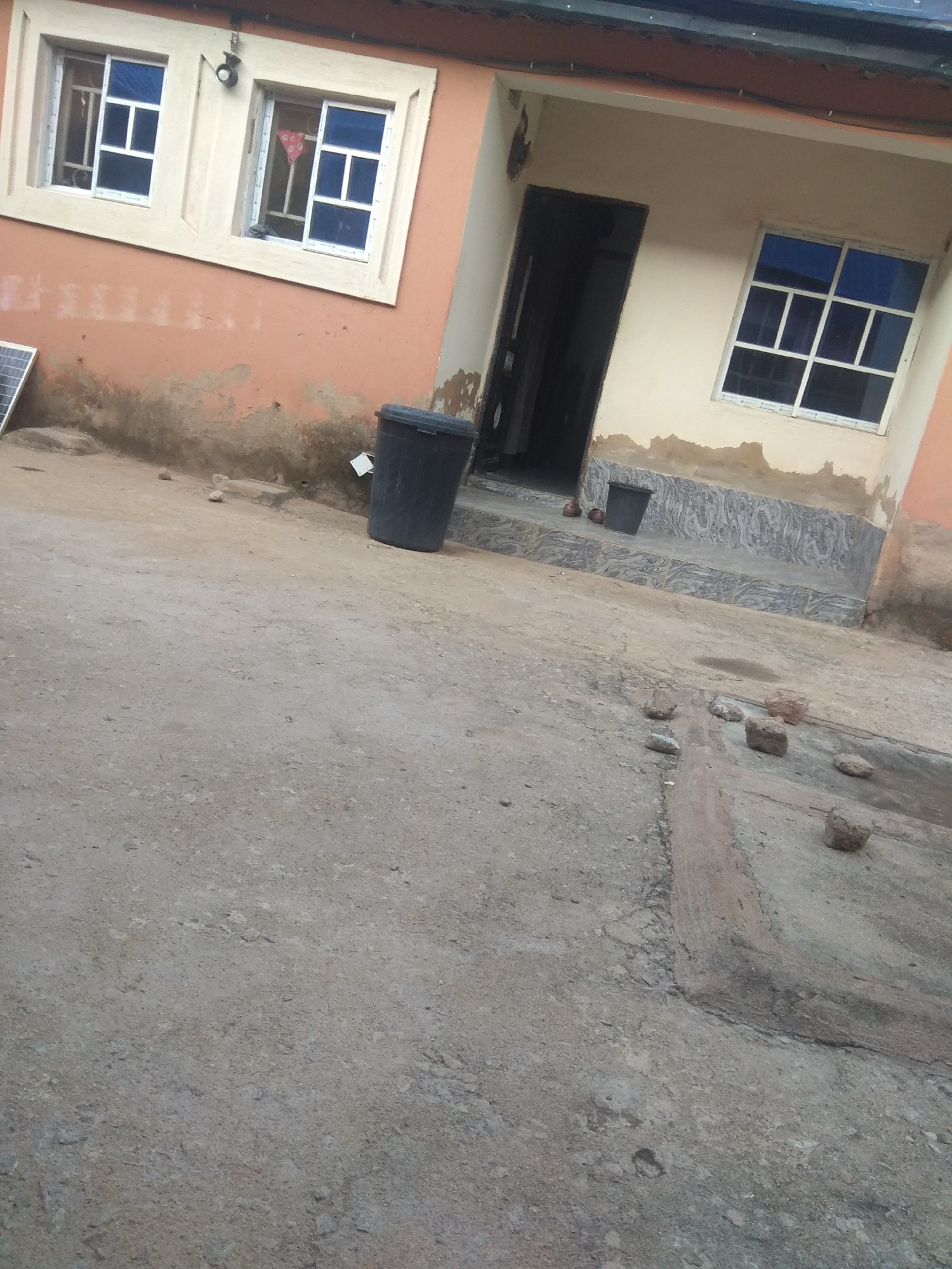 The image depicts an outdoor scene in Jalingo, Nigeria, focusing on the exterior of a building and the surrounding ground. The building's facade is painted in a peachy-orange color, with some areas showing signs of wear and tear. There are two windows visible on the upper part of the building, each with white frames and blue panes divided by white mullions. A security light is mounted above and between the windows. To the right of the windows, a dark doorway is visible, leading into the building.
The ground in front of the building is made of concrete or packed earth, showing cracks and an uneven surface. Several rocks are scattered on the ground, particularly in the foreground and middle ground. A large, dark plastic trash bin is situated to the left of the doorway, near the building's foundation. A solar panel is leaning against the building on the far left. The overall impression is of a somewhat weathered and utilitarian urban environment.