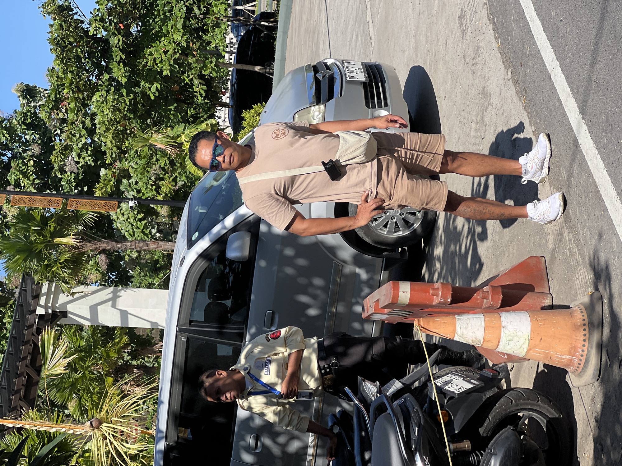 In a bright, sunlit outdoor scene in Denpasar, Indonesia, two men stand near a silver van. The man on the left, wearing a beige uniform with "SATPAM" visible on his shirt and a blue lanyard, stands with his left hand on a black scooter. He appears to be a security guard. The man on the right, wearing a beige t-shirt and tan shorts, sunglasses, and white sneakers, stands with his hands on his hips. He has a light-colored crossbody bag and a small black device attached to his shirt. In the foreground, an orange traffic cone with white and green stripes stands next to a red and white barrier. Palm trees and other lush greenery are visible in the background, casting dappled shadows on the ground. The road has a white painted line.