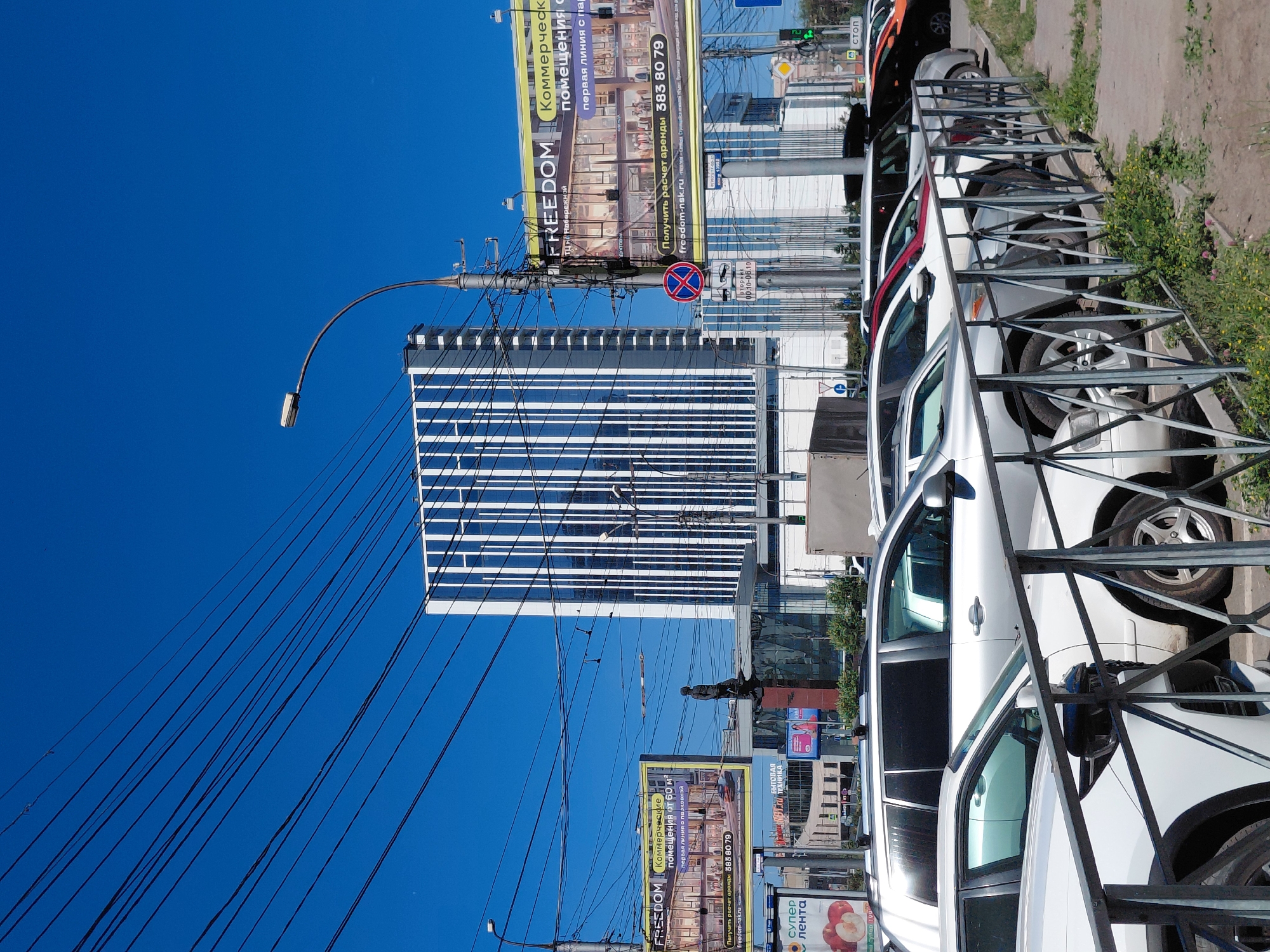 This is a vertical, eye-level, outdoor shot of a city street on a sunny day. The foreground features a row of white cars parked alongside a metal fence. Behind the cars, a modern glass-and-steel building with horizontal blue and white stripes dominates the middle ground. A street lamp with a curved arm extends from the left, its wires crisscrossing against a clear blue sky. Several large billboards are visible, one promoting "FREEDOM" for commercial spaces and another advertising "Супер лента" (Super Ribbon), possibly for a supermarket. Text on the billboards indicates "помещения от 60 м²" (premises from 60 m²) and a phone number. The scene suggests a commercial area with ongoing development or business activity. The time of day appears to be daytime, and the weather is clear and bright.