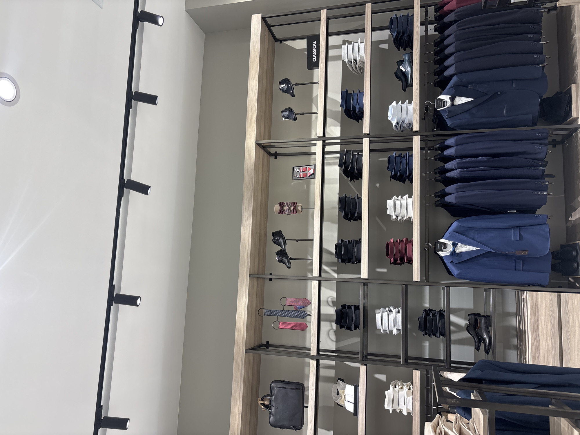 The image shows a retail store display of men's formal wear, likely a suit shop, located in Vratsa, Bulgaria. The display features racks of suits, shelves stocked with folded shirts and ties, and mannequins or stands showcasing shoes. The lighting appears to be overhead track lighting. There are no people visible in the image. The store environment is clean and organized, with a focus on showcasing the merchandise. The color palette is dominated by dark blues, grays, and whites.
