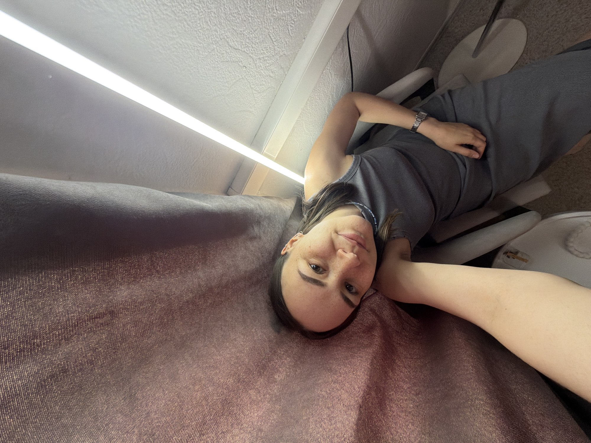 The image shows a woman lying on a brownish-purple velvet-like surface, possibly a couch or bed. She is wearing a grey tank top and matching pants or skirt, with a watch on her left wrist. Above her is a white ceiling with a fluorescent light fixture. To her right is a white chair and a stand with a circular base. The location is Chaikovsky, Russia.