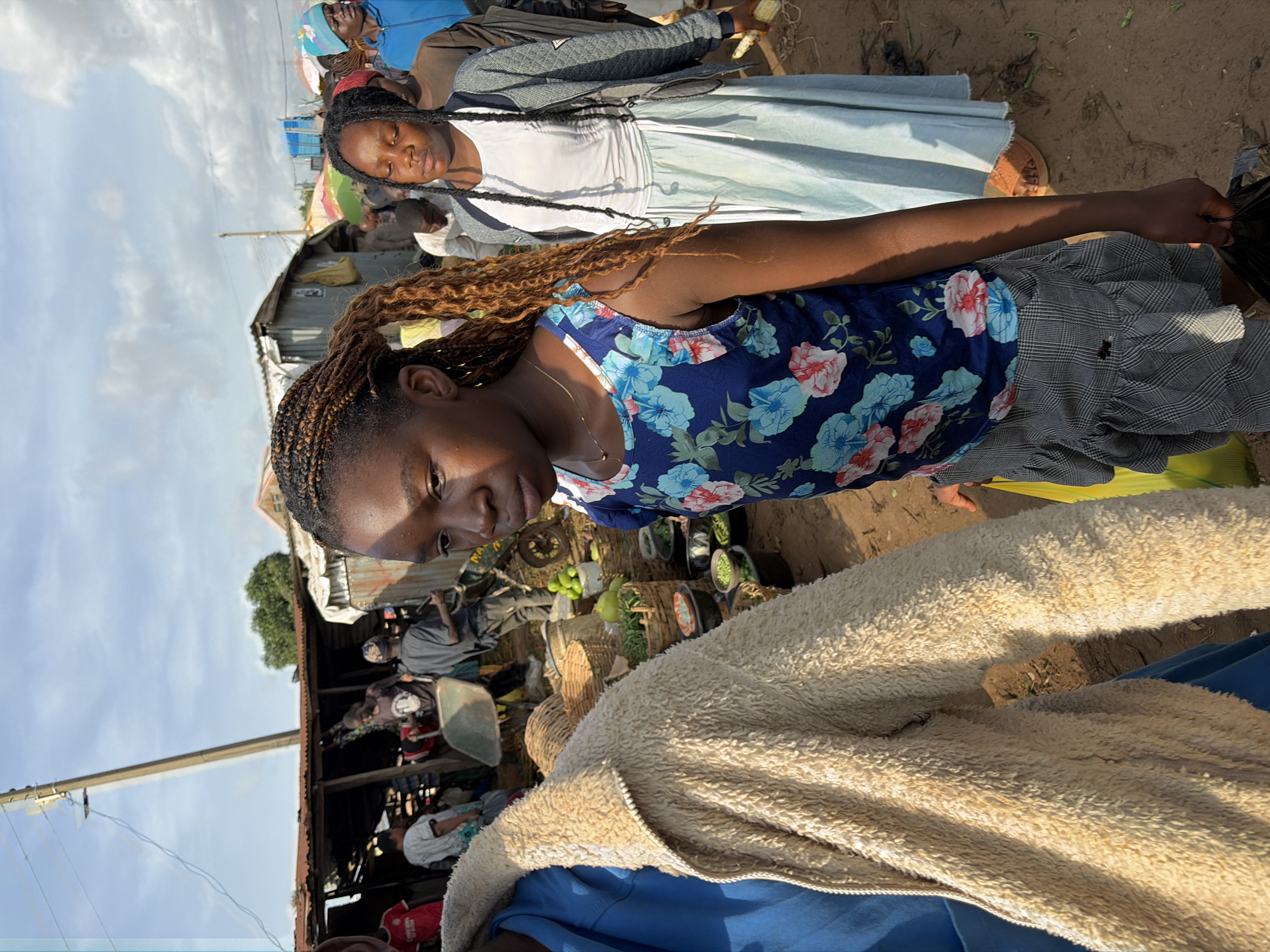 In Gura Top, Nigeria, a young girl with braided hair is captured in a medium shot, looking down and to the right. She wears a blue tank top adorned with a floral pattern and a grey and white checkered skirt. Her right hand holds a black plastic bag. To her left, another individual is partially visible, wearing a light beige fuzzy jacket over a blue garment. Further in the background, a bustling marketplace is evident with numerous people, market stalls with corrugated metal roofs, and various goods displayed, including produce in baskets and buckets. Some individuals appear to be engaged in shopping or working within the market. The environment suggests an outdoor setting with a dirt ground and a partly cloudy sky overhead.