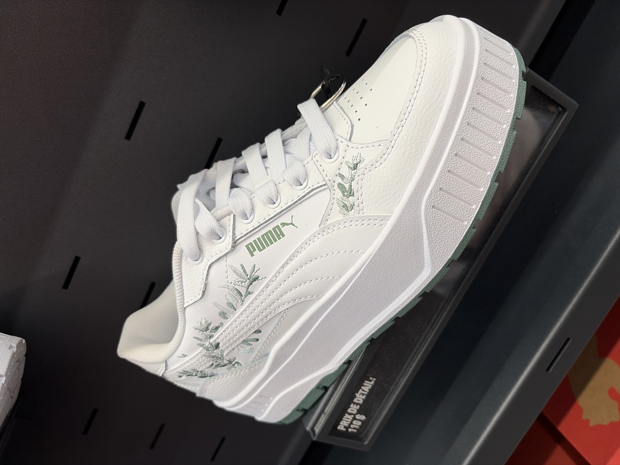 A white Puma sneaker with green floral embroidery and green accents on the sole is displayed on a slanted black shelf. The sneaker is positioned at an angle, with the laces visible. A price tag in French, reading "PRIX DE DETAIL: 110 $", is attached to the shelf below the shoe. The background is a dark, textured surface with several horizontal black slots. The lighting is bright, highlighting the shoe and the price tag. The image appears to be taken in a retail environment, possibly a shoe store. There are no people or other animals visible in the image.