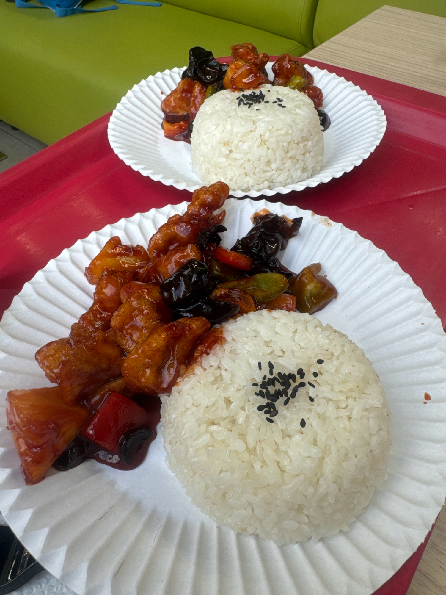 The image shows two paper plates of food, each containing rice and a stir-fry dish, placed on a red surface. The food appears to be Asian cuisine, possibly sweet and sour chicken or pork, with pieces of meat, vegetables, and a glossy sauce. The rice is white and fluffy, topped with black sesame seeds. The plates are white and have a pleated design. In the background, a portion of a green sofa or chair is visible, along with a light-colored wooden surface. The lighting suggests an indoor setting, possibly a restaurant or food court. There are no people visible in the image. The image does not contain any information that would allow for its location to be identified as Irkutsk, Russia.