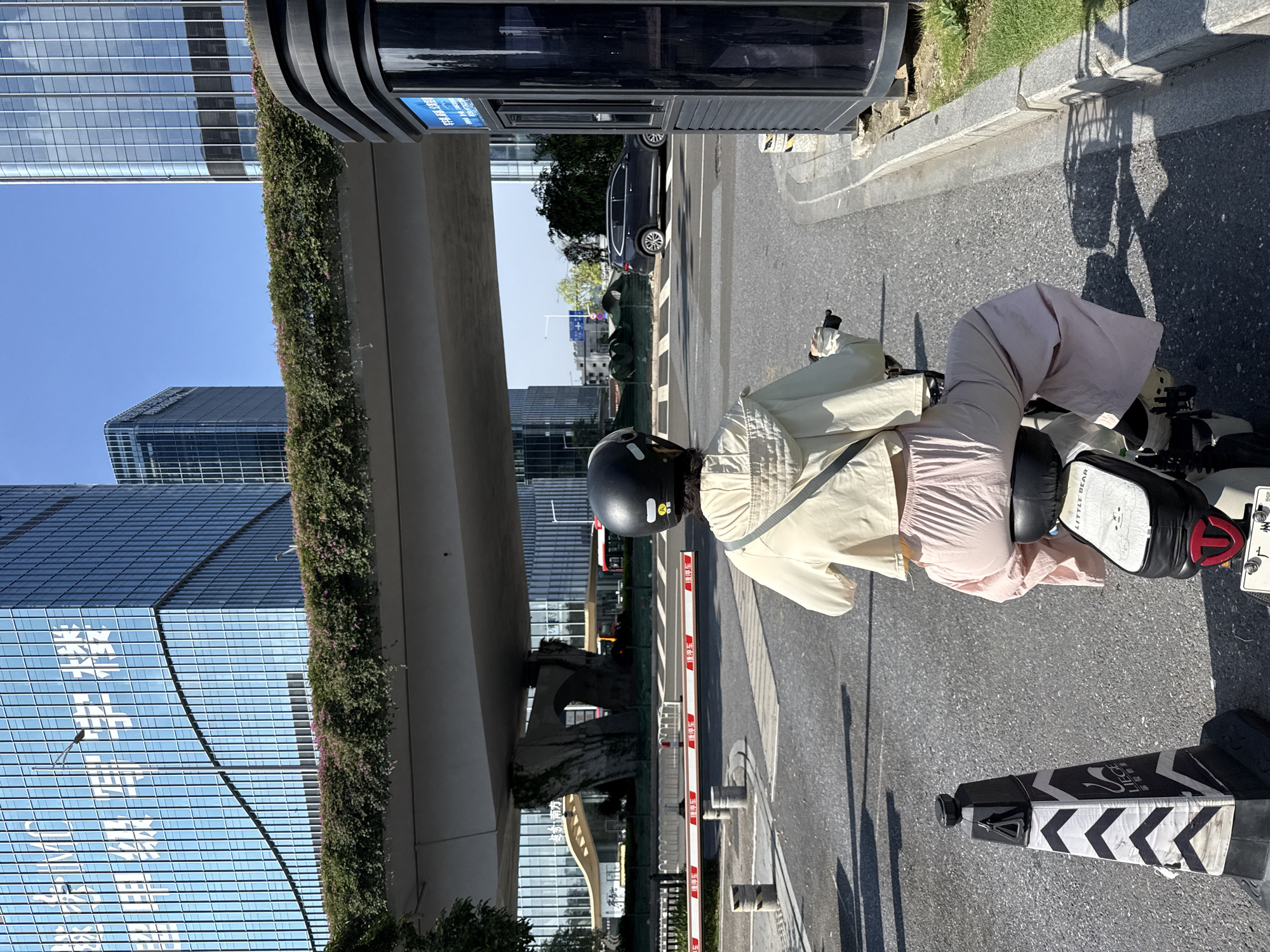 A person rides an electric scooter under a bridge in Huangpu District, China. The person wears a light yellow jacket, pink pants, and a black helmet. The scooter has a white seat with a cartoon bear graphic and "LITTLE BEAR" text. The scene is set in a modern urban environment with tall glass buildings, a lush green overpass, and a blue sky. A black vehicle and a red bus are visible in the background, along with a parking barrier and traffic cones.