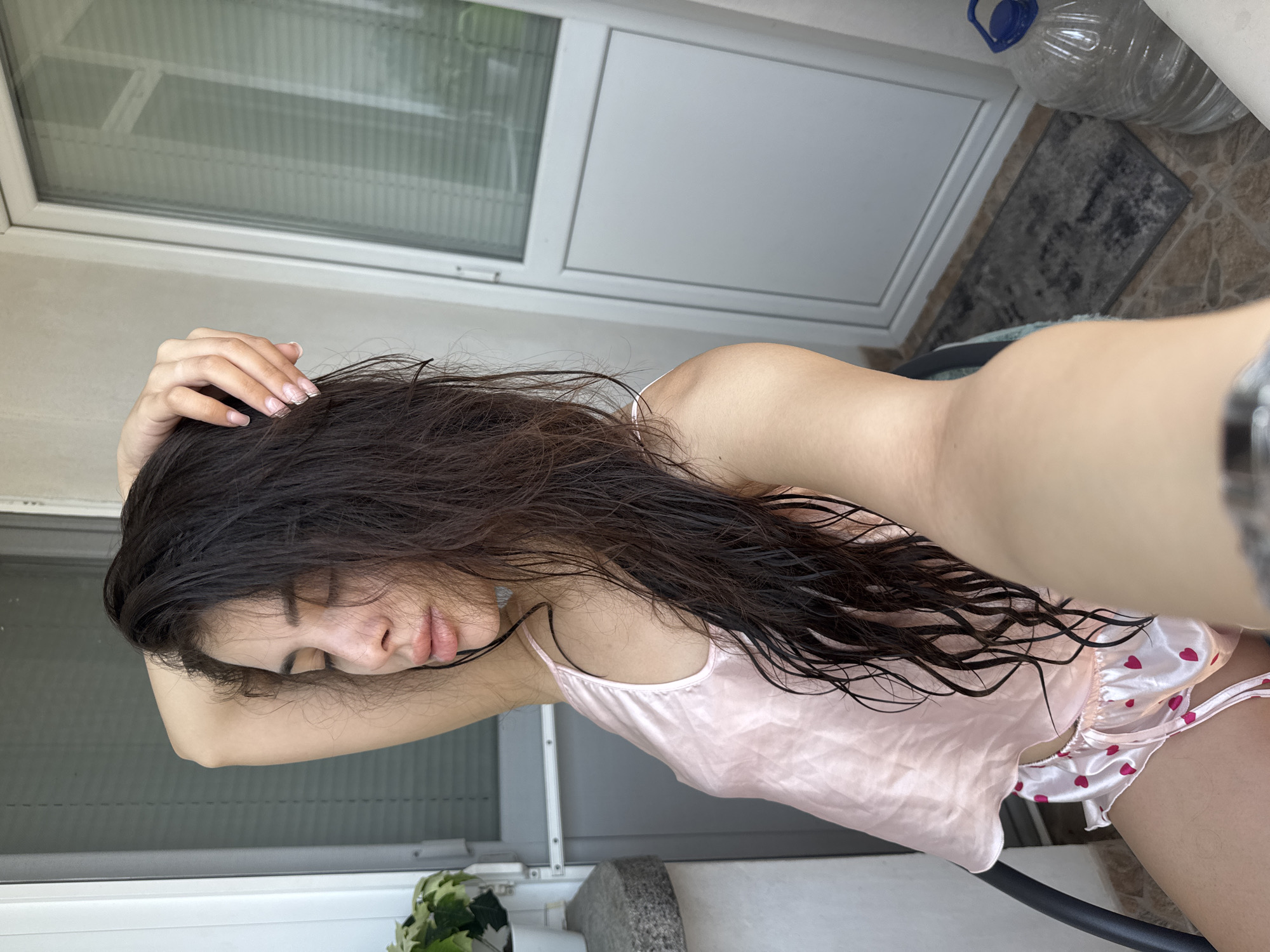 A young woman is captured taking a selfie on what appears to be a balcony or room in Plovdiv, Bulgaria. She is seated or leaning, with her right arm raised and hand resting on the back of her head, her eyes closed, and lips slightly parted. Her long, dark brown hair is visibly wet and falls over her shoulders and chest. She is wearing light pink satin pajamas, consisting of a camisole top with thin straps and shorts patterned with red hearts. Her fingernails exhibit a light French manicure style. The background features a white PVC door with a glass panel, through which horizontal blinds are visible. To the left, a small potted plant sits on a stone ledge. The floor is tiled, and a small dark mat lies in front of the door. A large, clear plastic water bottle with a blue handle and cap is situated on the floor to the right of the door.