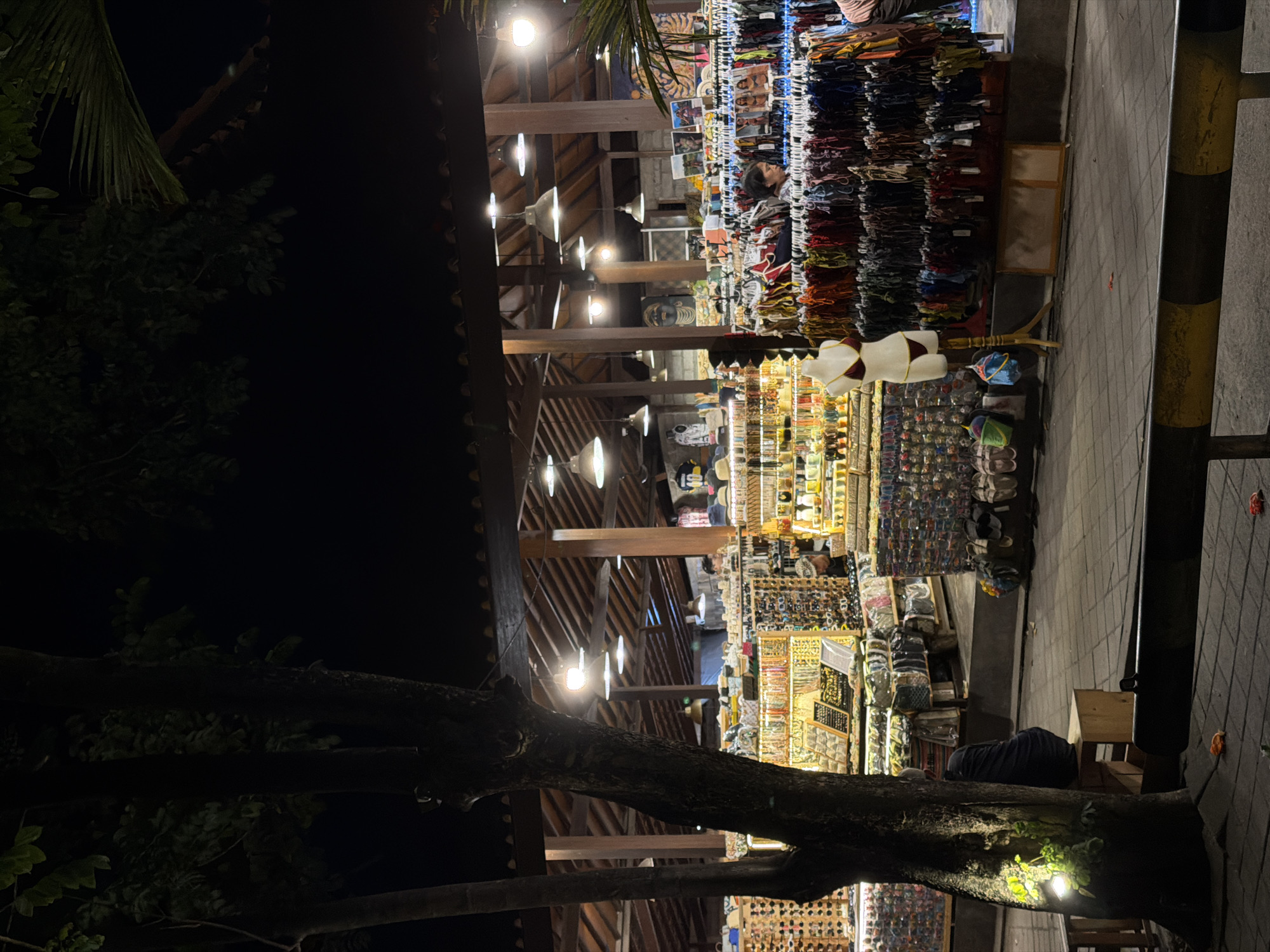The image captures a night scene in North Kuta, Indonesia. The photograph depicts an outdoor market stall illuminated by hanging lights. A variety of items are on display, including clothing, jewelry, and souvenirs. A vendor can be seen behind one of the stalls. The photograph also shows a tree in the foreground with branches extending into the scene. A pedestrian walkway is visible to the right of the stalls. The environment is dark, with the market area providing the main source of light.