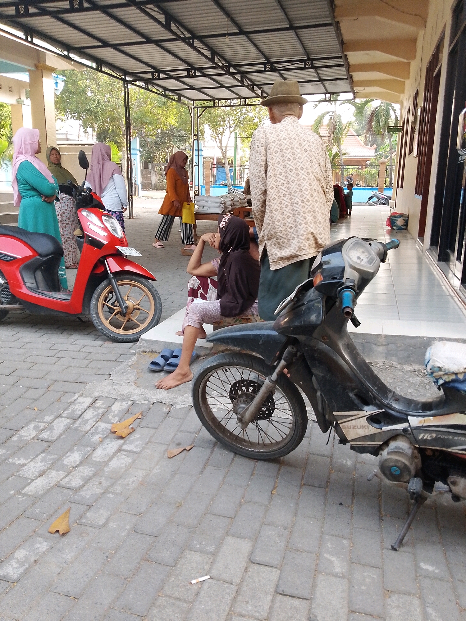 A rear view of an elderly man wearing a patterned shirt and a hat stands in the foreground, facing away from the camera. He is on a paved walkway. To his left, a red scooter is parked. Further left, three women in hijabs stand near a building entrance. One woman wears a teal hijab, another a green hijab, and a third a light pink hijab. Behind them, a building with a covered walkway is visible. To the right of the man, a black motorcycle is parked. A young girl in a headscarf sits on the ground near the motorcycle, facing the man. The environment appears to be outdoors, with trees and other buildings visible in the background. The ground is paved with interlocking bricks. The scene is likely in Wadang, Indonesia.