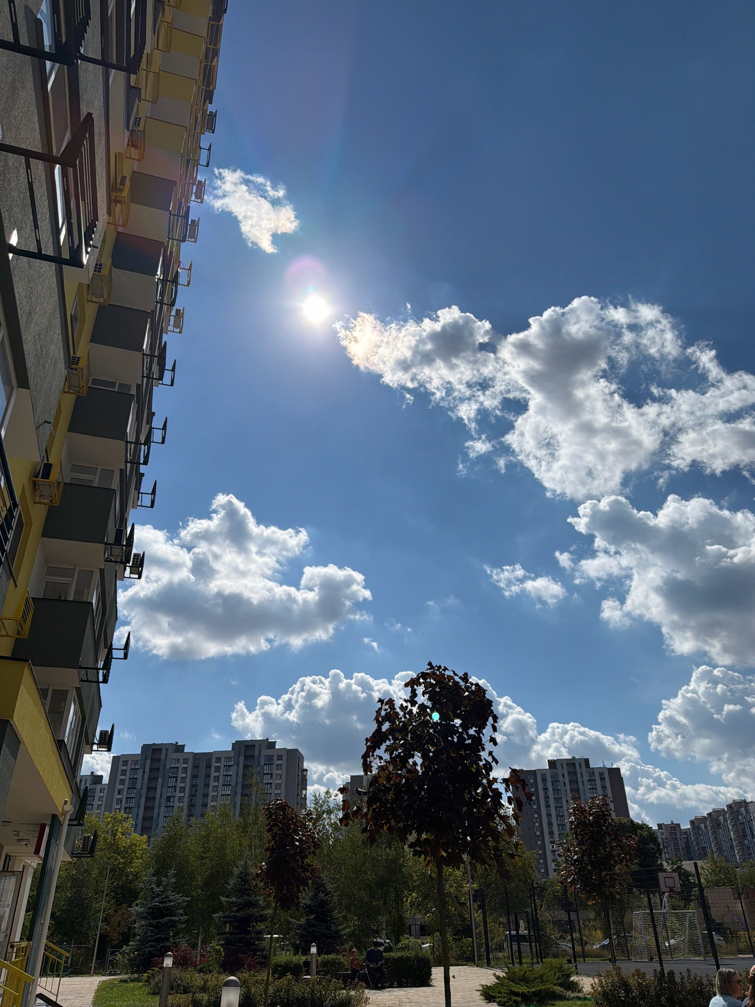 The image captures a sunny day in a residential area of Kyiv, Ukraine. A modern apartment building with yellow and gray facades dominates the left side of the frame, its balconies adorned with air conditioning units. The sky is a vibrant blue, dotted with fluffy white cumulus clouds and illuminated by the bright sun, which creates lens flare. In the mid-ground, a row of multi-story apartment buildings is visible behind a lush green treeline, suggesting a park or communal green space. Several trees, including one with dark red foliage in the foreground, add to the natural element. The ground level features a paved walkway, manicured bushes, and outdoor lighting fixtures. In the far right, a sports court with a basketball hoop and a soccer goal can be seen, along with a few people. The overall impression is of a well-maintained urban environment on a clear, pleasant day.