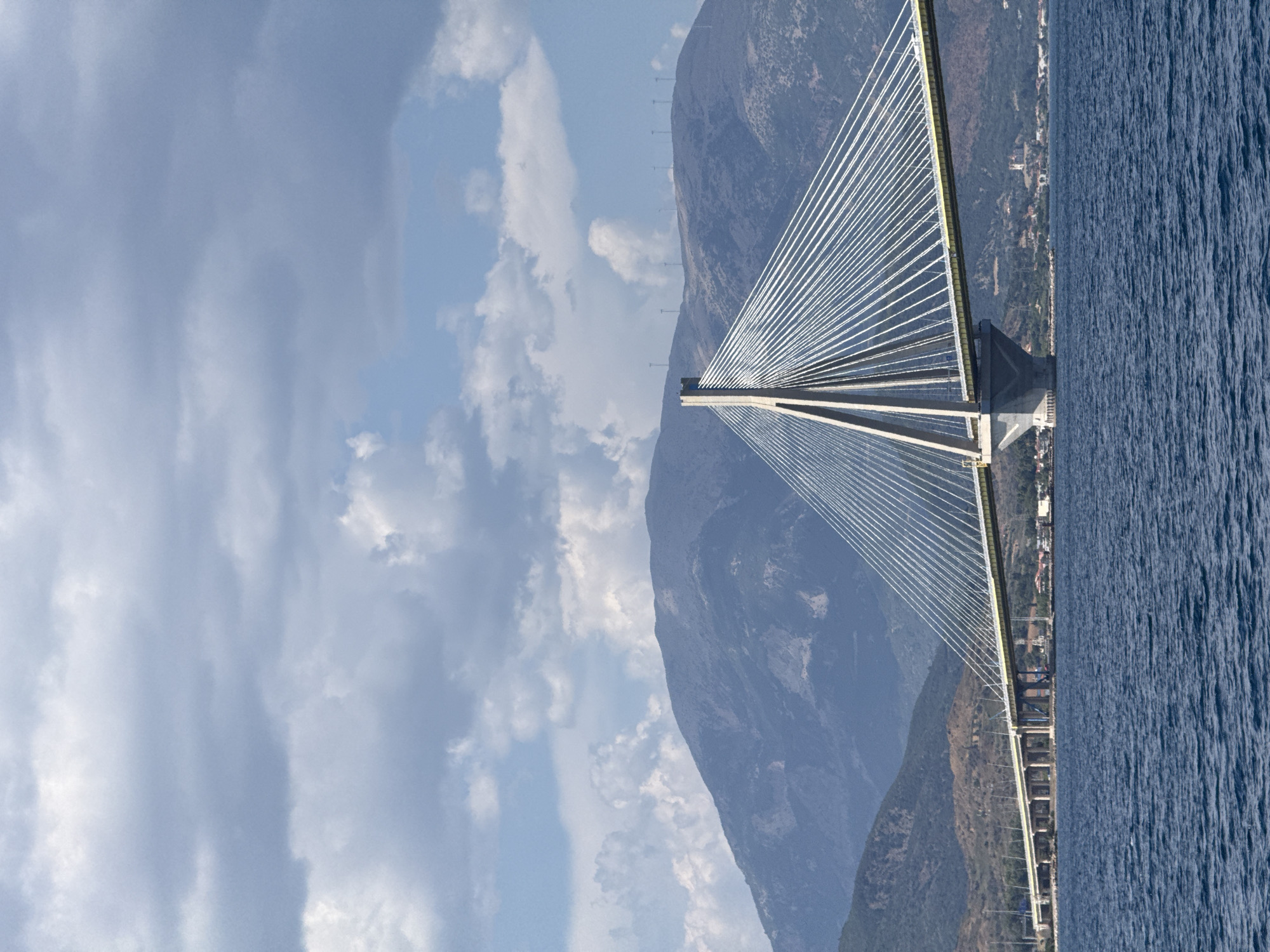This media file depicts a cable-stayed bridge spanning a body of water, likely a strait or bay, with a mountainous coastline on one side and the open sea on the other. The bridge is the dominant object, with its tall, slender pylons and numerous cables creating a striking geometric pattern against the sky and mountains. The sea is a deep blue with a rippled texture, suggesting it is somewhat rough or windy.
The scene is set during the day, under a partly cloudy sky with patches of blue visible between the white and gray clouds. The mountains in the background are rugged and covered in sparse vegetation, with a small settlement visible along the coastline. Several tall, thin structures, possibly electricity pylons or wind turbines, are lined up along the crest of the mountain.
The perspective is from a viewpoint slightly above the water, looking towards the bridge and the coastline. The image's orientation is rotated, showing the scene sideways. No people are visible in the frame, and no text is present. The overall impression is one of impressive engineering set against a dramatic natural landscape. The location context indicates this is likely the Rio-Antirrio Bridge in the Municipal Unit of Rio, Greece, which connects the Peloponnese peninsula with mainland Greece over the Gulf of Corinth.