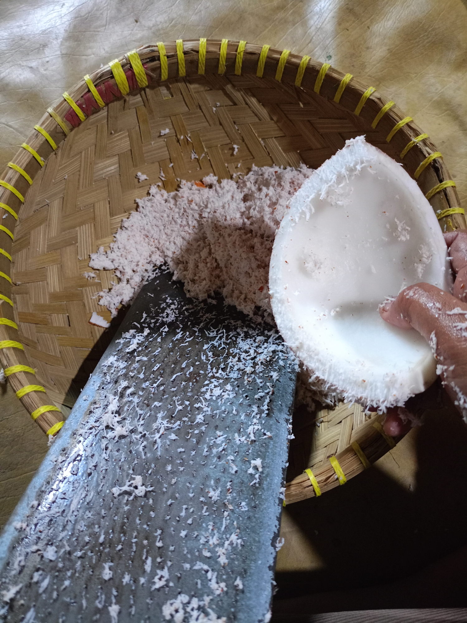 An overhead, close-up shot shows a person's hand grating coconut over a traditional woven bamboo tray. The tray is circular with yellow string binding its perimeter and appears to be resting on a textured, light brown surface. A large metal grater, covered in white coconut shavings, occupies the bottom left of the frame. To the right of the grater, a pile of grated coconut has accumulated. In the upper right quadrant of the tray, a half-coconut shell, also lined with white flesh and shavings, is being held by a hand. The scene captures the process of preparing grated coconut, likely for culinary use, in Jelbuk, Indonesia.