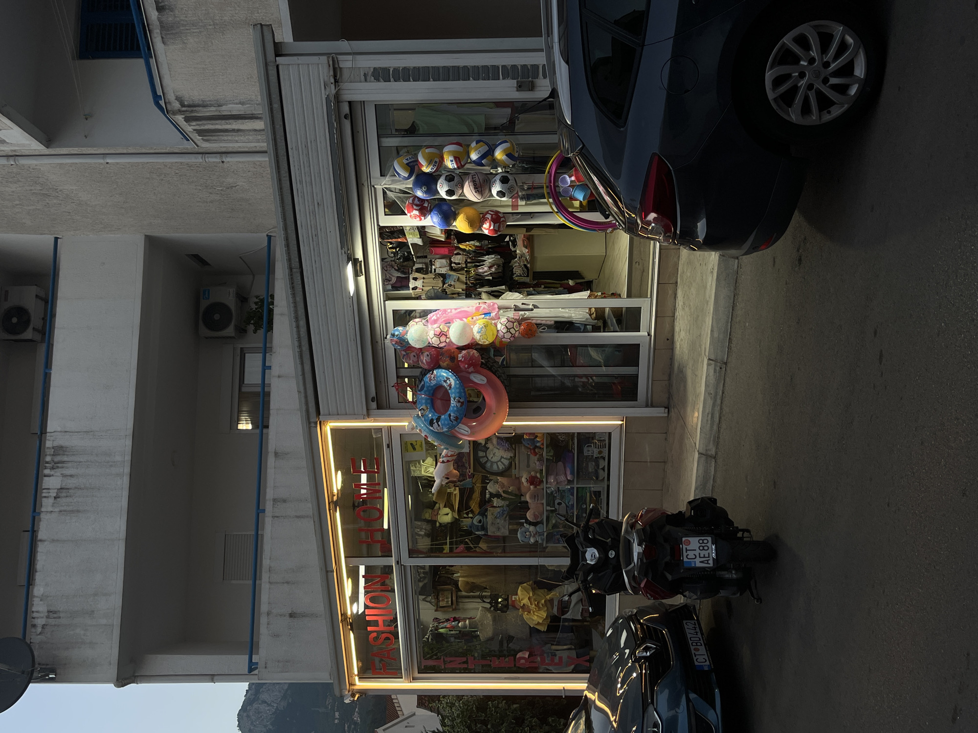 In the city of Budva, Montenegro, at ground level, a retail establishment with illuminated signage reading "FASHION HOME" is visible. The store's interior displays various items, including clothing on mannequins, stuffed toys, and household goods. Outside the store, inflatable rings and an assortment of balls are displayed. In front of the store, parked on the asphalt street, are a blue car and a red scooter. The scooter is positioned directly in front of the store's entrance. A dark gray car is parked to the right of the scooter, obscuring part of the store's facade. The buildings in the background appear to be residential or commercial, with balconies and air conditioning units visible. A mountain range is discernible in the distance, under a pale sky.