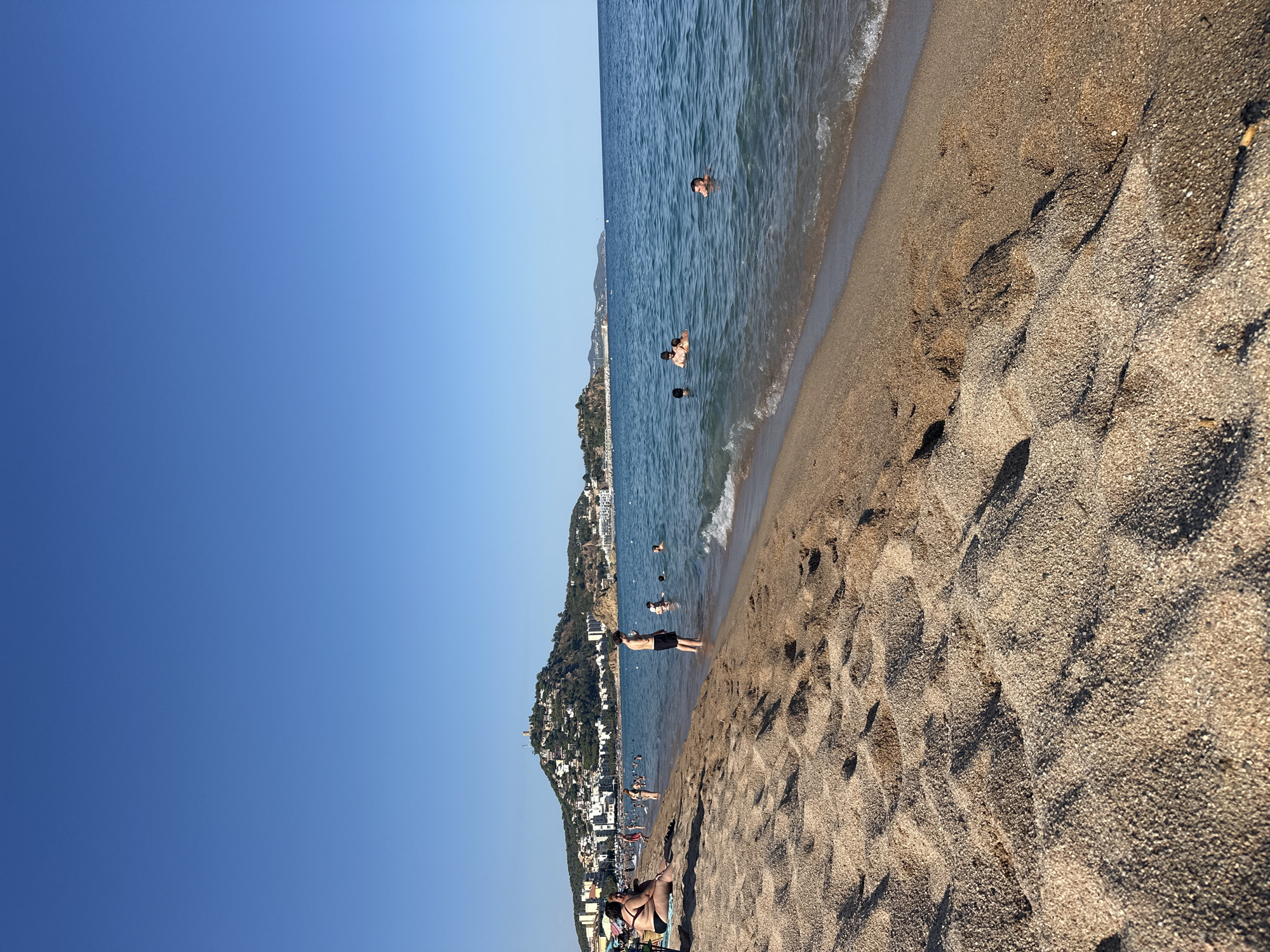 A wide-angle, eye-level, daytime shot captures a sandy beach in Blanes, Spain, with the Mediterranean Sea stretching towards the horizon. In the foreground, the sandy shore is textured with ripples and footprints. The sea, a clear blue, laps at the shore with gentle waves. Several people are visible in the water, swimming and wading. On the beach, individuals are sunbathing and standing near the water's edge. In the middle ground, a coastal town is nestled against a verdant hill dotted with buildings and topped with a prominent structure, likely a lighthouse or church. The sky above is a bright, cloudless blue.