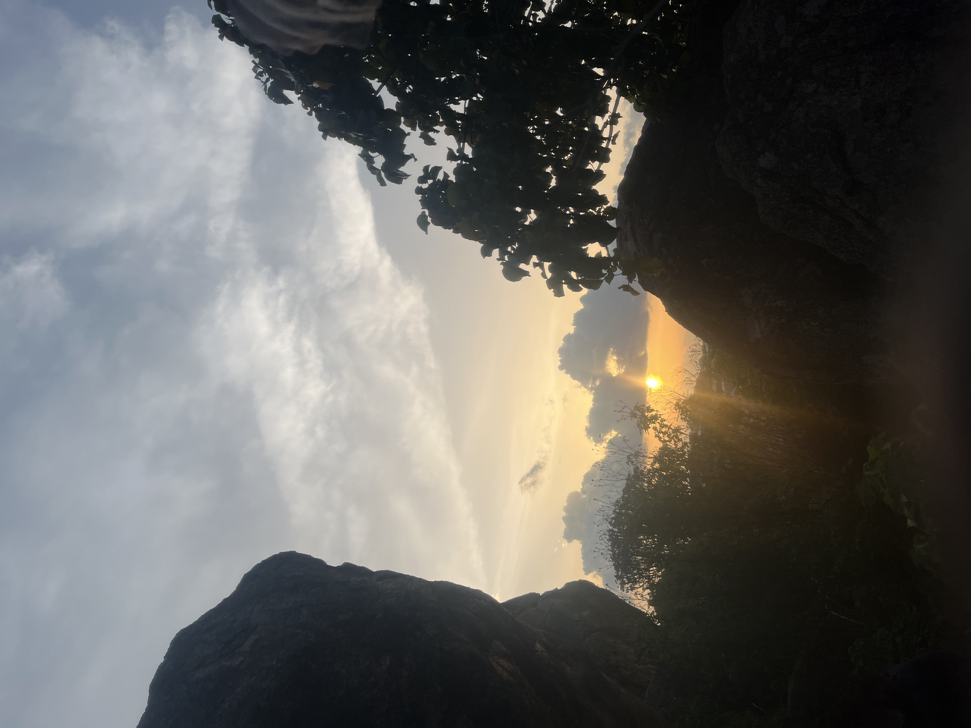 This image depicts a natural outdoor landscape in Barakin Akawo, Nigeria, captured during either dawn or dusk. The scene is dominated by silhouetted natural elements against a warm, cloudy sky.
The upper portion of the frame features a partly cloudy sky, transitioning from cooler grey tones at the top to warmer orange and yellow hues towards the horizon. The sun is visible as a bright, golden orb in the lower center-right, partially obscured by clouds and casting prominent light rays downwards and to the left.
The left side of the frame is occupied by a large, dark, irregular rock formation or cliff face, rendered in deep silhouette against the brighter sky. The upper right portion features a dense, dark tree with visible leaves and branches, also silhouetted. Below this tree, another large, dark rock formation is discernible. The lower half of the image is filled with dark, dense foliage, including bushes and smaller trees, all in deep shadow, obscuring the ground plane which appears to be rocky and vegetated. No human figures, animals, or man-made structures are present or discernible within the frame.