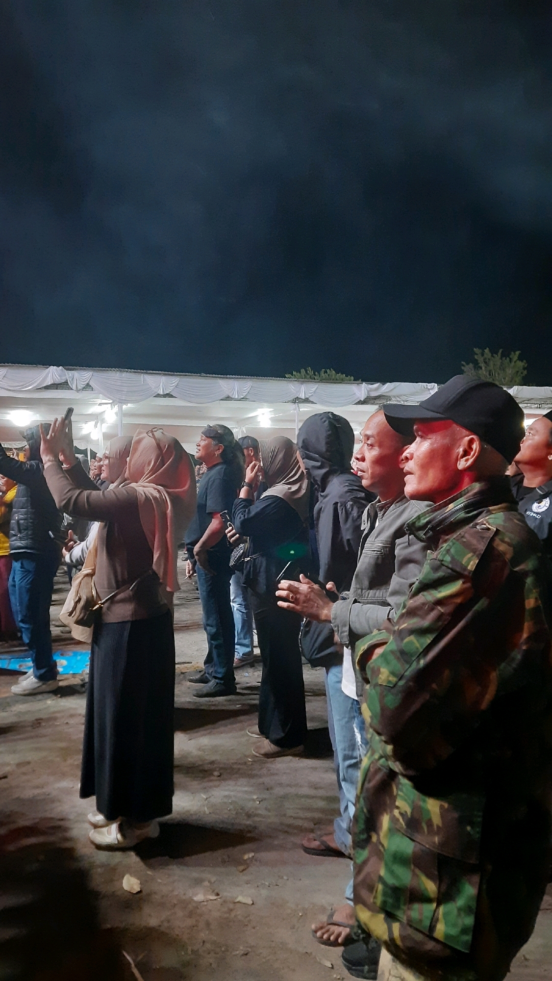 The image depicts a gathering of people at night, likely an outdoor event, with a white tent structure in the background. The foreground shows a man in camouflage clothing and a black baseball cap, facing right. To his left, a group of people are visible, some wearing hijabs, and others in casual clothing. Several individuals are holding up mobile phones, suggesting they are recording or taking pictures. The ground appears to be dirt or gravel. The scene suggests a public event or concert in Minggir, Indonesia.