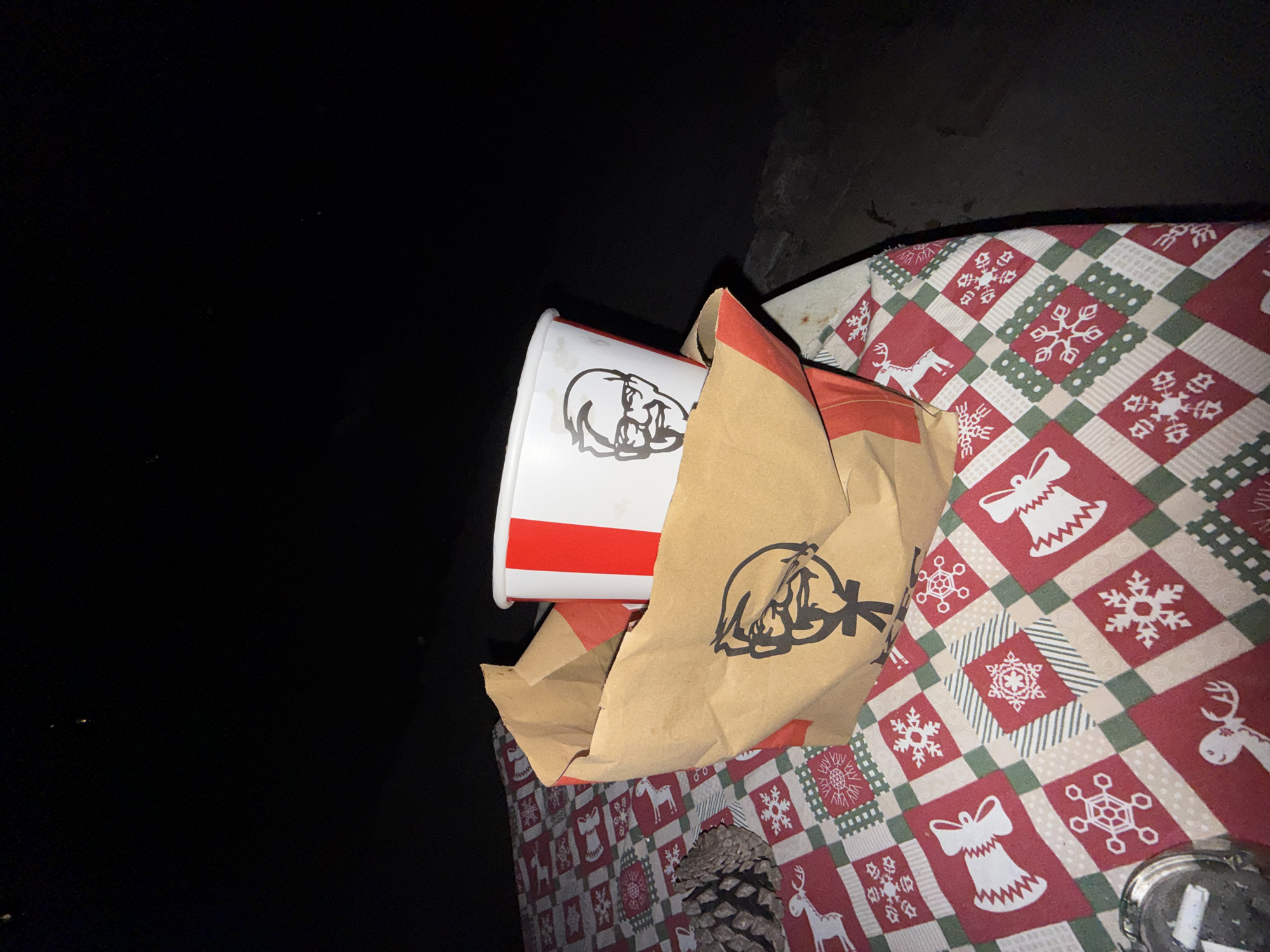 The image depicts a nighttime scene in Varna, Bulgaria, presenting a table covered with a festive, holiday-themed tablecloth. The tablecloth displays a checkered pattern primarily in red, white, and green, adorned with motifs including snowflakes, bells, and reindeer. Centrally positioned on the table is a crumpled brown paper KFC bag, from which a white KFC bucket partially emerges. Both the bag and the bucket feature the black outline logo of Colonel Sanders; the bucket also displays prominent red vertical stripes. To the immediate left of the KFC bag, a dark brown pinecone rests on the tablecloth. In the lower-left foreground, a circular metallic ashtray containing cigarette ash and a visible cigarette butt is present. The background is characterized by profound darkness, with only minimal, indistinct glimmers or shapes at its furthest extents, suggesting an outdoor or dimly lit environment. No individuals are depicted in the image.
