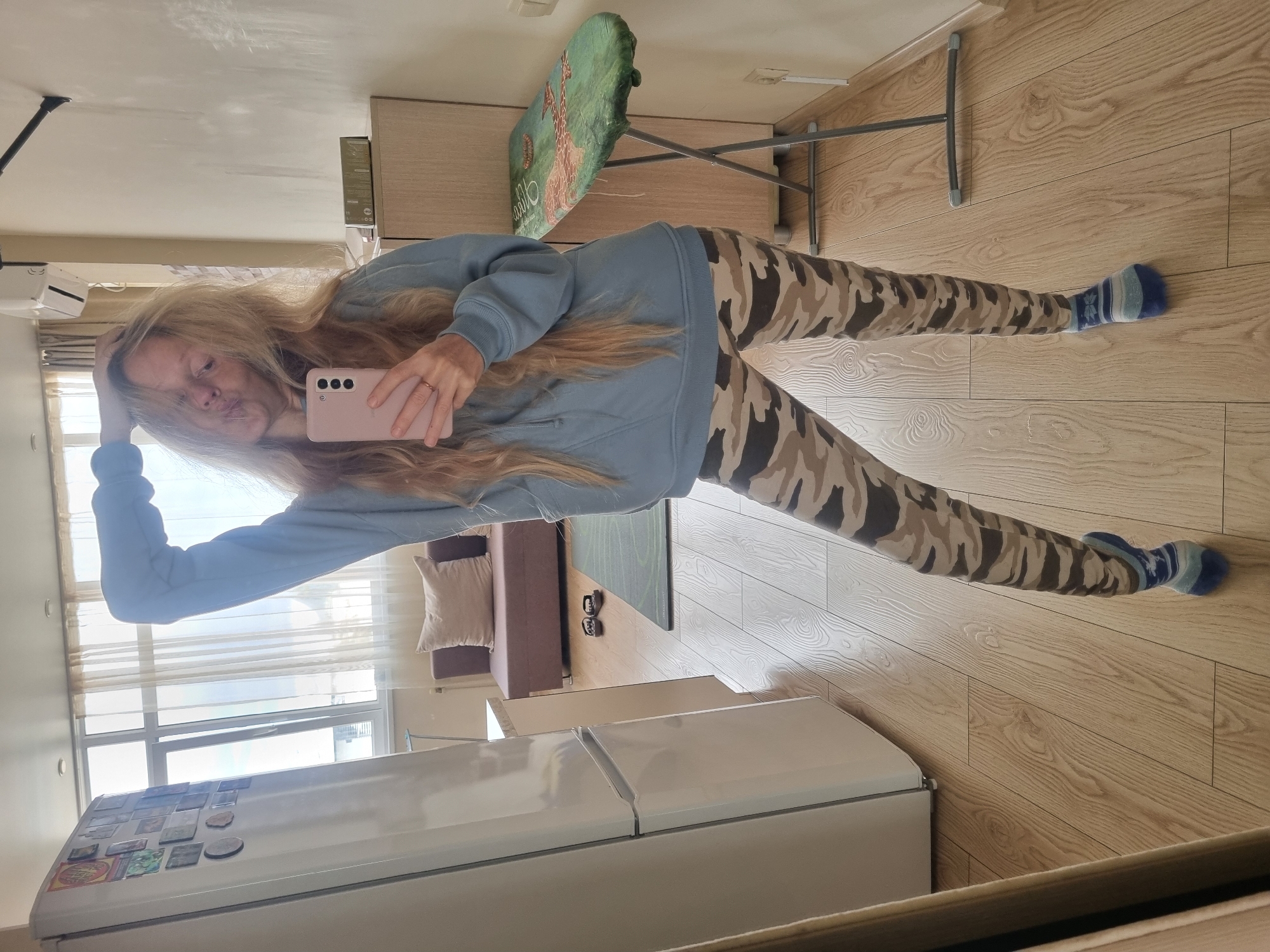 A full-length selfie of a blonde woman in a light blue sweatshirt and camouflage-patterned leggings is taken in what appears to be an apartment. The woman is holding a pink smartphone in her right hand, using it to capture her reflection in a mirror. Her left hand is resting on her head. Her legs are spread wide, and she is wearing blue and white striped socks.
The background shows a typical apartment interior with laminate flooring. A white refrigerator is in the foreground, along with a glimpse of a brown couch with a pillow. Further back, there is a green ironing board and other furniture that appears to be part of a living space. The presence of a magnet-covered refrigerator, a couch, and an ironing board suggests this is a lived-in space.
The image is taken indoors, likely during the daytime, given the natural light coming through the window. The location, Sochi, Russia, is provided but cannot be verified based on the visual elements of the image alone.