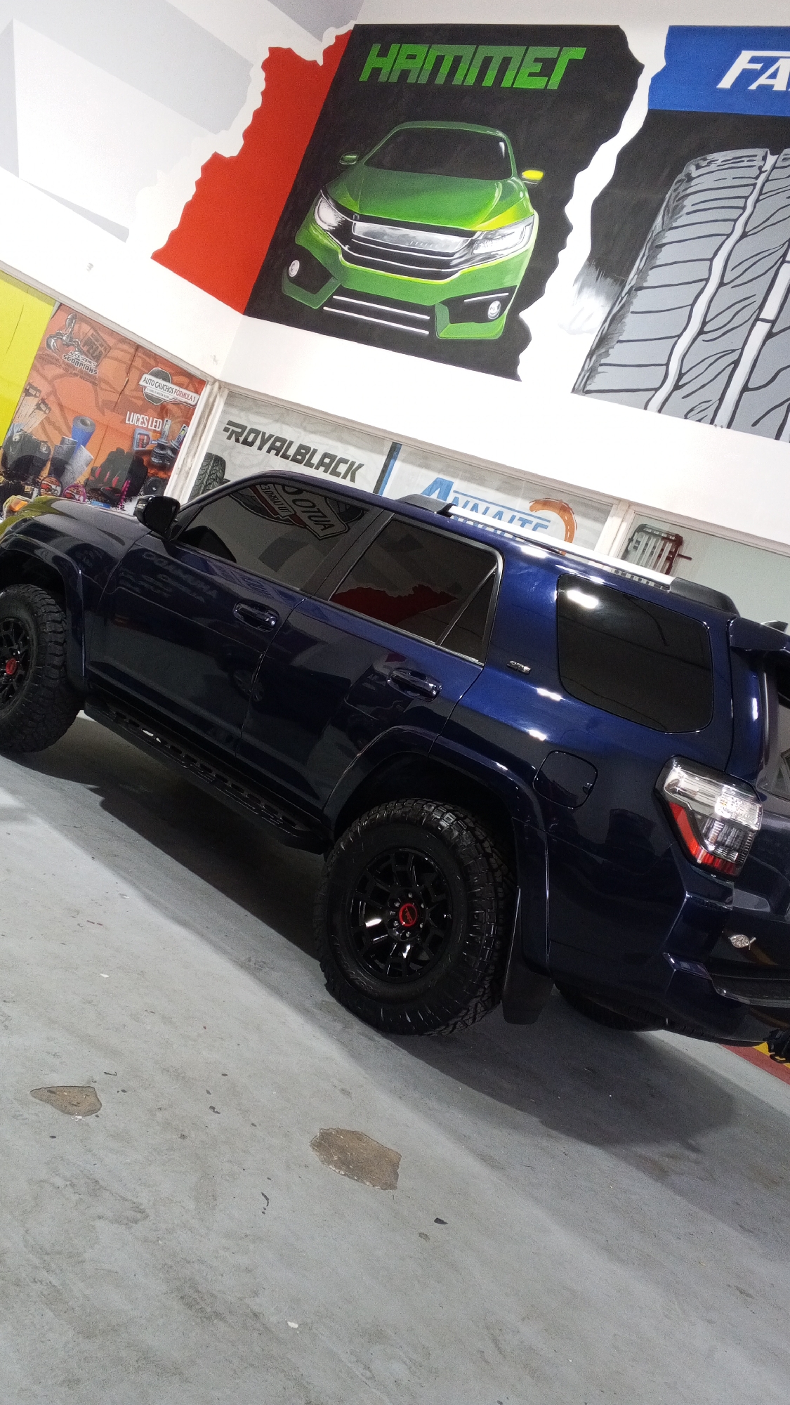 An eye-level shot captures a dark blue, possibly custom, SUV parked indoors, likely within an auto shop or showroom. The vehicle, resembling a Toyota 4Runner, is positioned slightly angled towards the right, showcasing its left side. It features black multi-spoke wheels with red center caps, rugged all-terrain tires, black running boards, and heavily tinted windows. A small fish emblem is visible on the rear bumper.
The background consists of brightly painted walls adorned with large-scale murals. To the upper left, a mural depicts a vibrant green car, similar to a modified Honda Civic, beneath the bold green text "HAMMER." This section has a black background bordered by abstract white and red shapes. Adjacent to it on the right, another mural features a detailed tire tread pattern with the partially visible blue text "FA" above it, against a blue background, separated from the car mural by a jagged white line.
Reflected in the SUV's windows and visible on a wall in the mid-ground is the brand name "ROYALBLACK." Other banners and signs on the left wall are partially visible, displaying text such as "AUTO CAUCHOS FORMULA Y" (Auto Tires Formula Y), "LUCES LED" (LED Lights), and "SCORPIONS." The floor is a light gray concrete, showing some irregular dark stains or wet patches. The lighting appears artificial, suggesting an indoor setting.