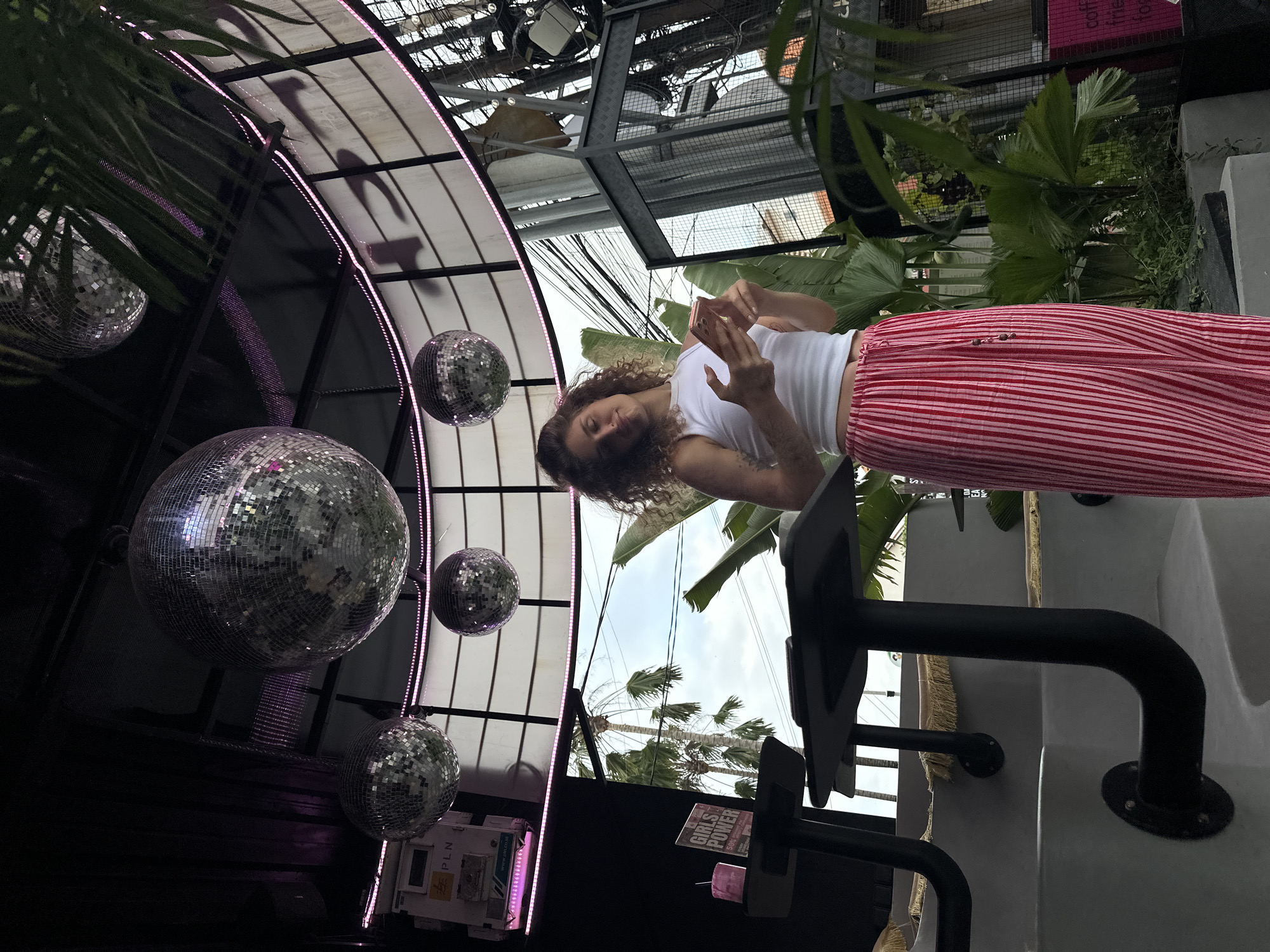 A woman with curly brown hair, wearing a white tank top and red and white striped pants, is looking down at her phone. She is standing in an outdoor setting, possibly a bar or restaurant, with disco balls hanging overhead. The scene is illuminated by bright, natural light, suggesting it is daytime. Lush green foliage, including palm trees, is visible in the background, consistent with a tropical location like North Kuta, Indonesia. Wires and metal structures are also part of the overhead architecture, adding an industrial touch. A sign that says "GIRLS POWER" is visible on the right side of the frame. The overall atmosphere appears relaxed and casual.