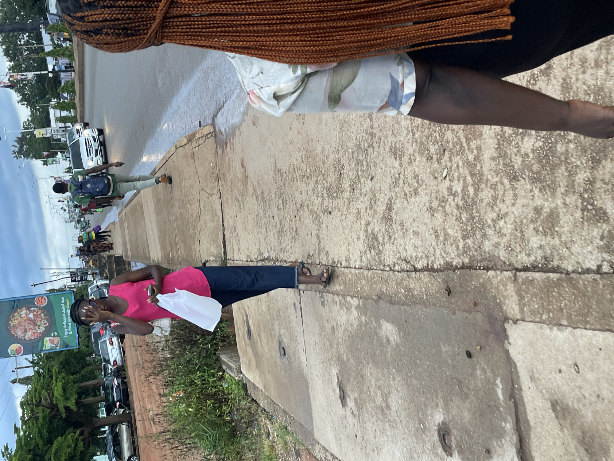 The image captures a street scene in Jos, Nigeria, on a cloudy day. In the foreground, the back of a woman with long, braided, dark red hair is visible, wearing a patterned shirt and dark pants. To her left, a woman in a bright pink tank top and dark pants walks on a concrete sidewalk, holding a white plastic bag and a mobile phone. She is partially shielding her face with her hand and wearing dark sunglasses. Behind her, a young man with a backpack walks further down the sidewalk, away from the camera. The sidewalk runs parallel to a paved road where several vehicles, including a white Mercedes-Benz, are driving. The environment includes green trees, streetlights, and a large billboard advertising Jollof rice. The ground appears wet in some areas, suggesting recent rain.