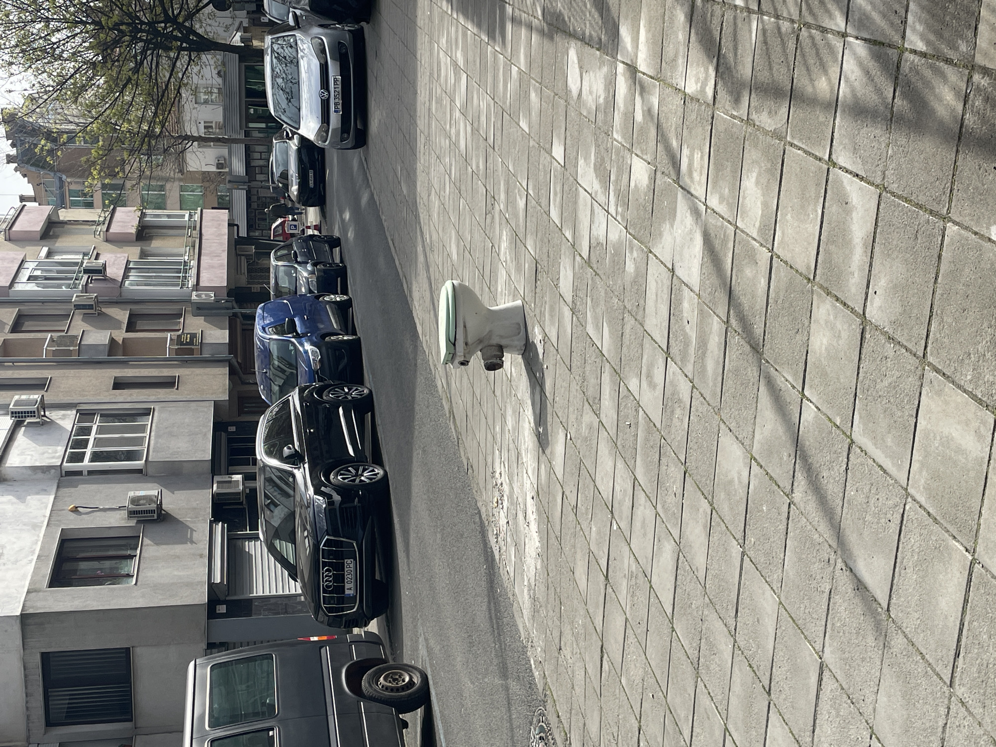 The image depicts a street in Burgas, Bulgaria, lined with parked cars and buildings. A toilet stands on the sidewalk. The day appears bright.